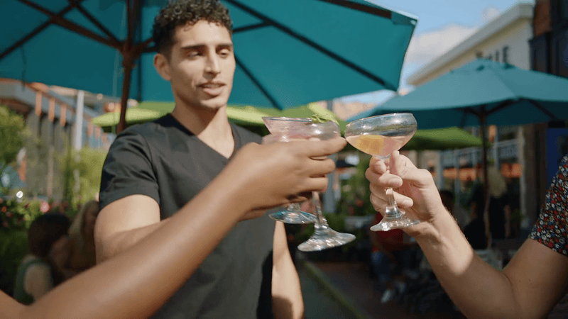 A group of friends making a toast with colorful cocktails under patio umbrellas on an outdoor restaurant deck in Grand Rapids.