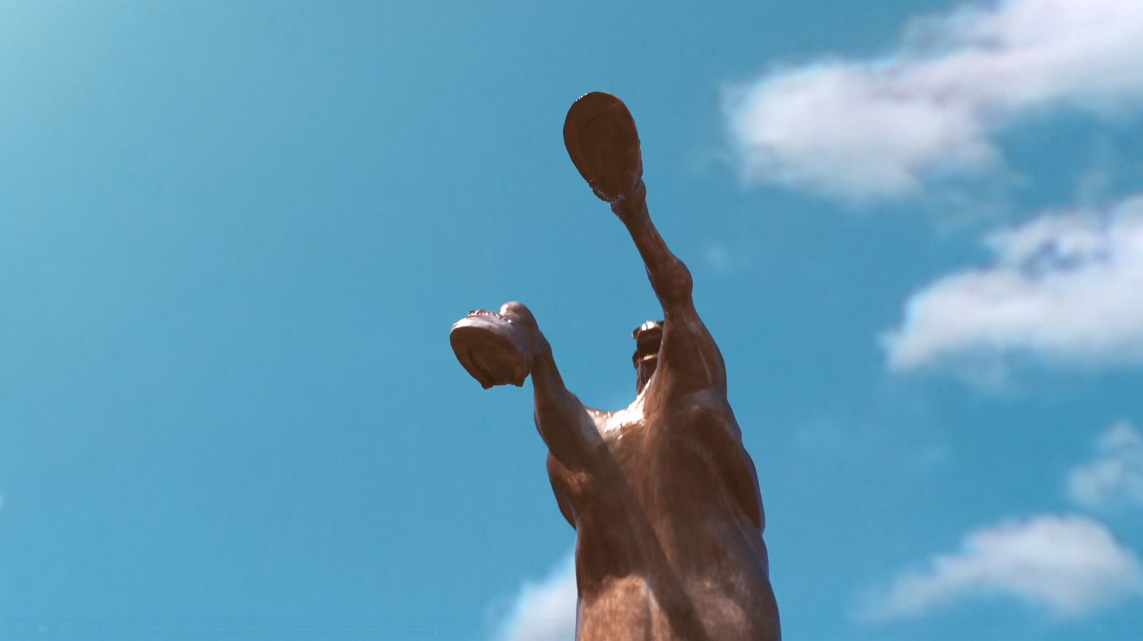 dramatic low-angle view looking straight up past the rearing front hooves of the bronze horse sculpture into a bright blue sky with white clouds.