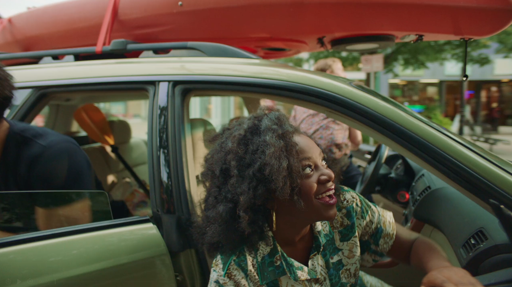 A smiling woman looks out the passenger side window of an SUV loaded with a red kayak on its roof rack.