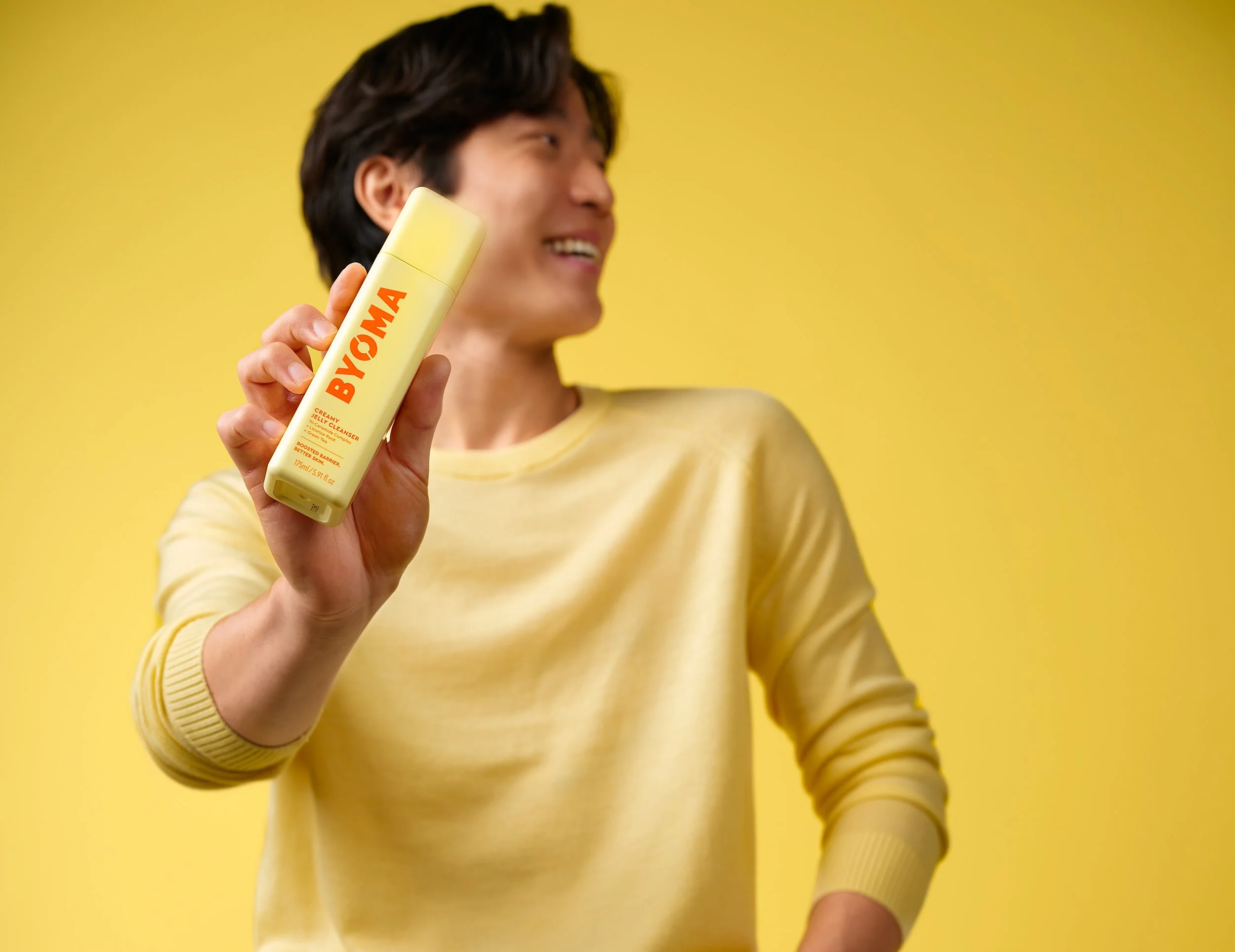 Model holding Byoma skincare product against a yellow background.