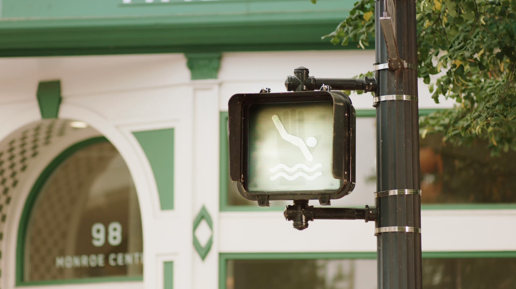 A crosswalk signal outside a storefront labeled 98 Monroe Center showing an animated glowing swimmer icon instead of a walking figure.