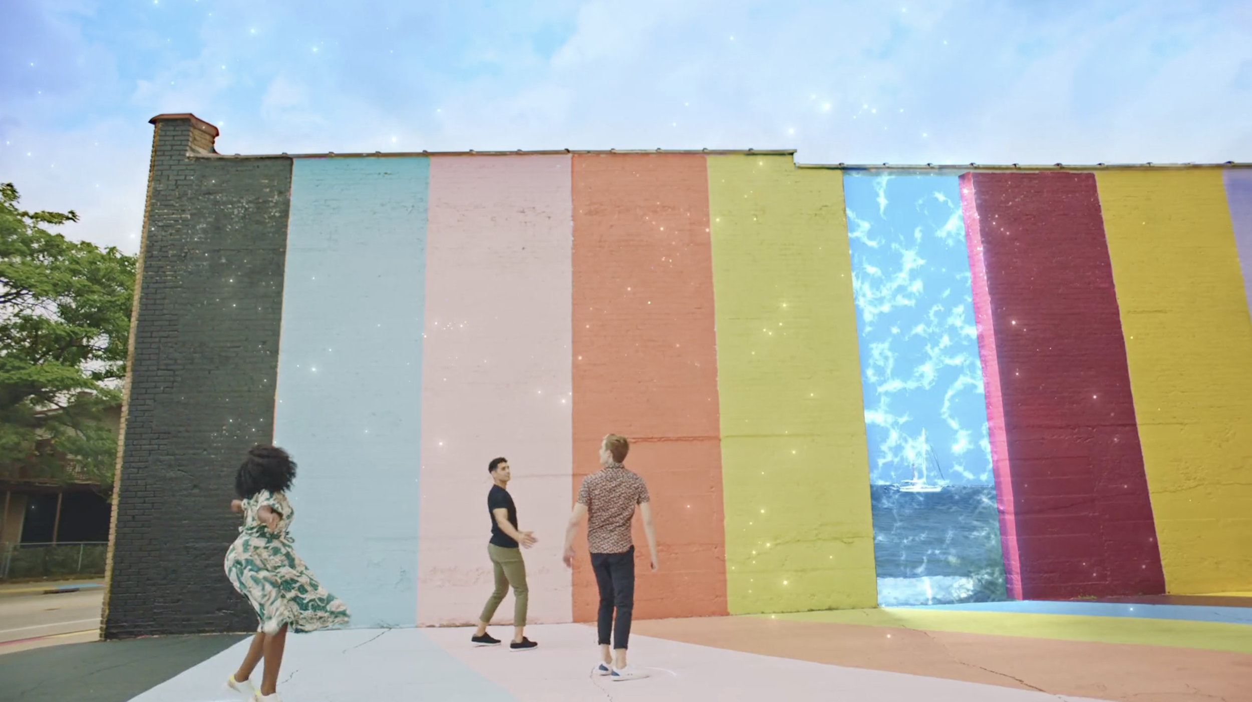 Three people walking past a large outdoor rainbow-striped wall mural with magical sparkling animations and a glowing blue panel effect.