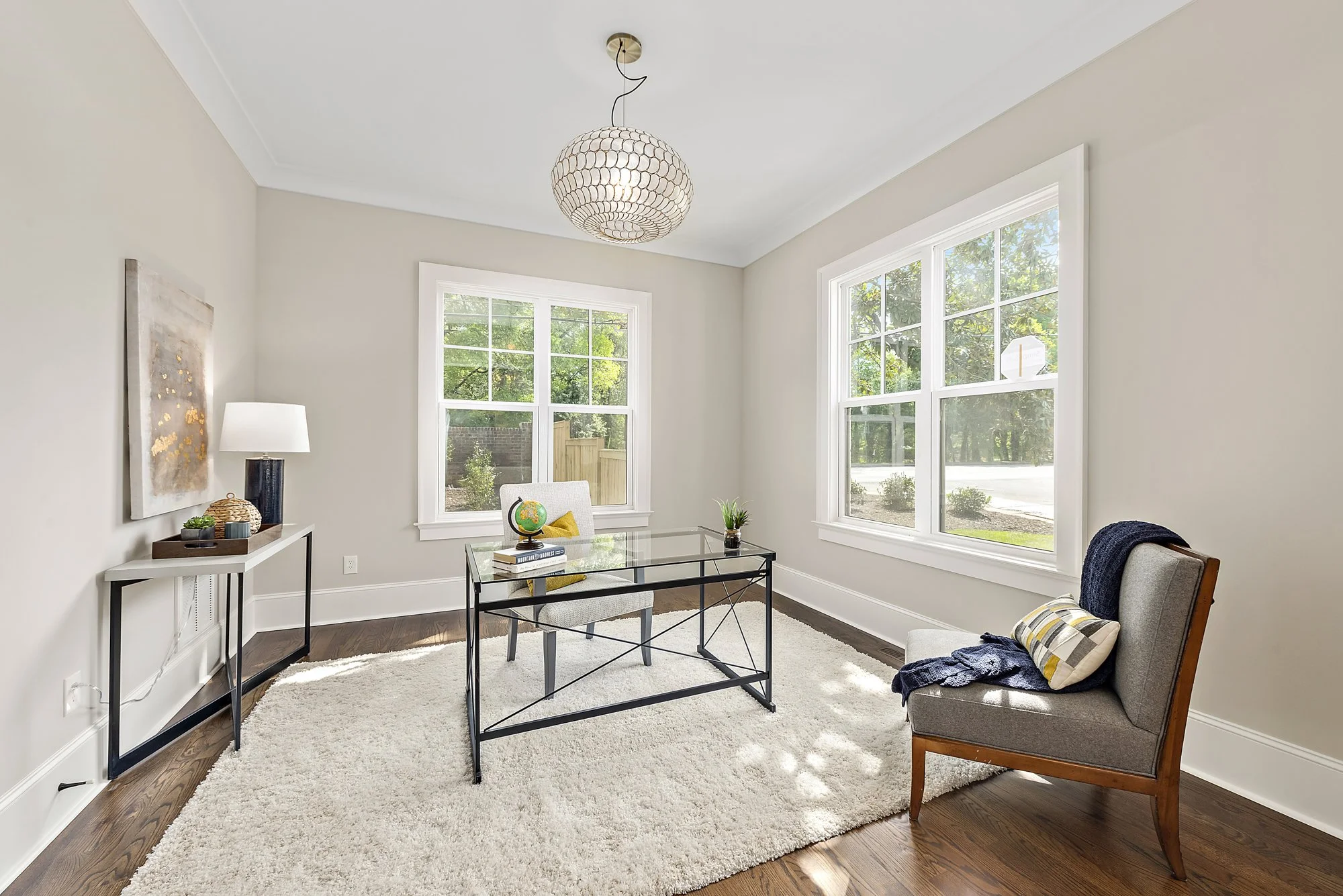 Bright home office with a glass-top desk, modern chair, large window, and a rug on hardwood floor. Decor includes a plant, globe, and lamp.