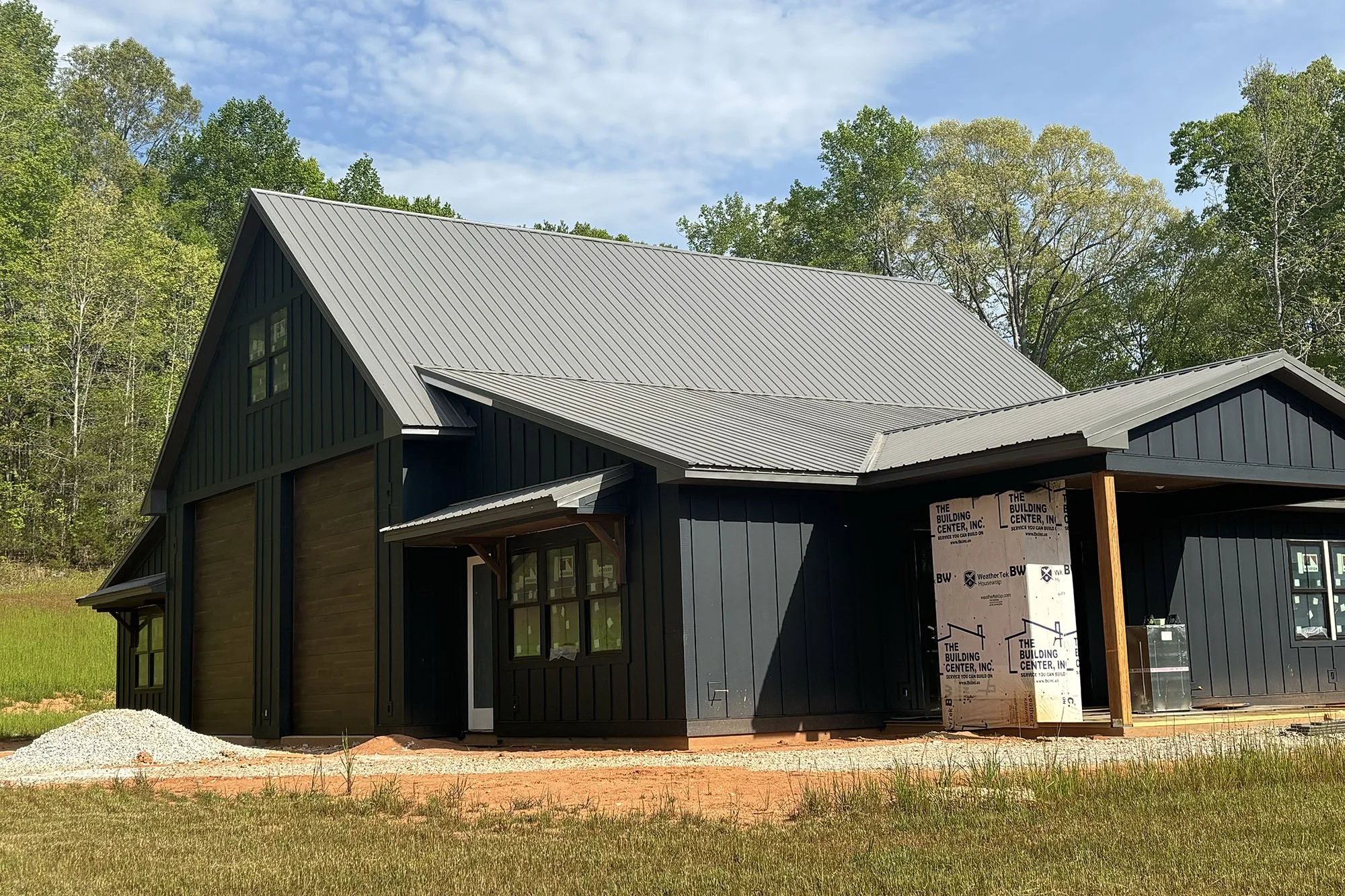 A two-story house under construction with dark blue vertical siding and a metal roof, set against a backdrop of trees and a partly cloudy sky.