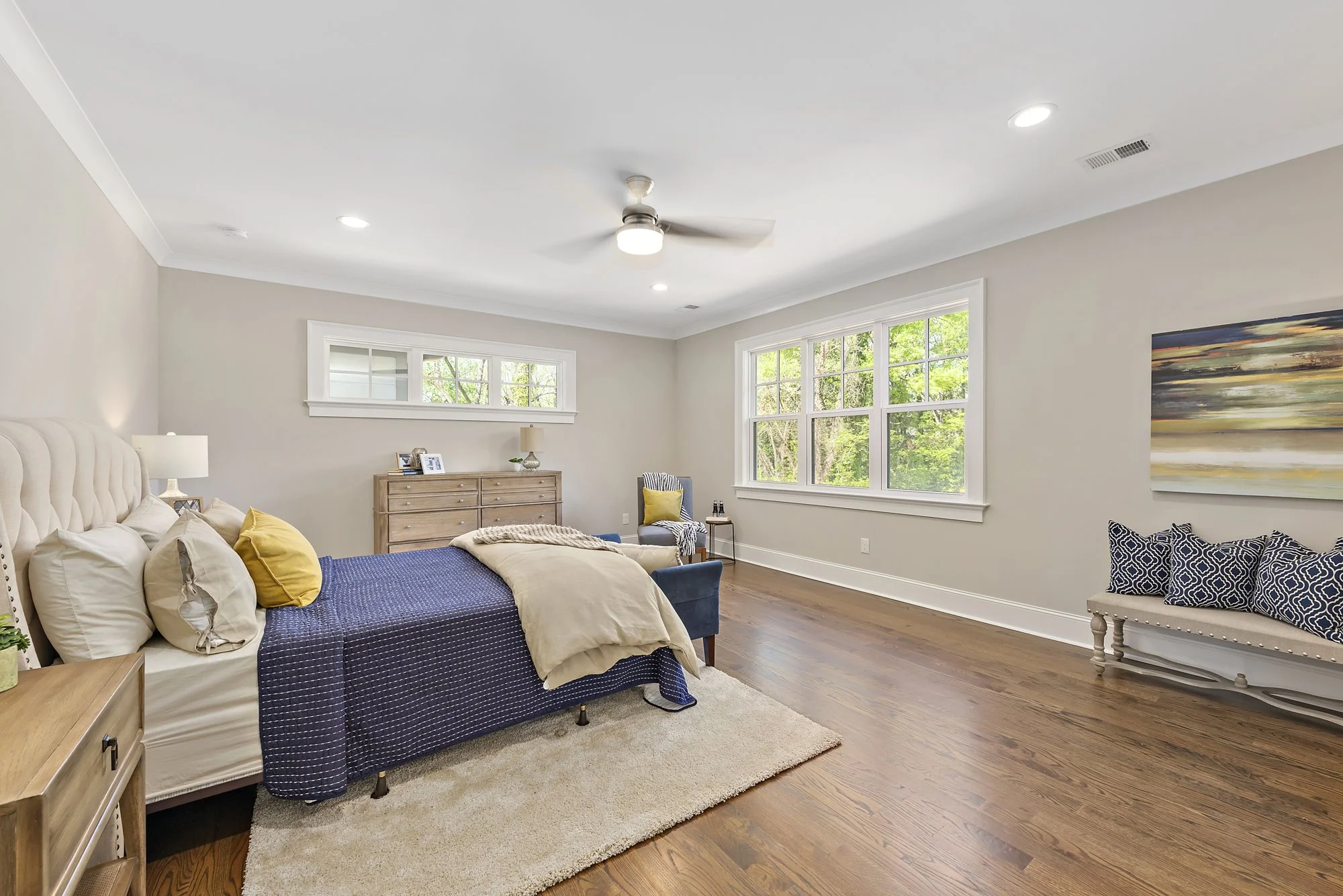 Spacious modern bedroom with wooden floor, featuring a bed with blue and beige linens, a ceiling fan, large windows with a view of greenery, a cushioned bench, a dresser with lamps, and decorative pillows.