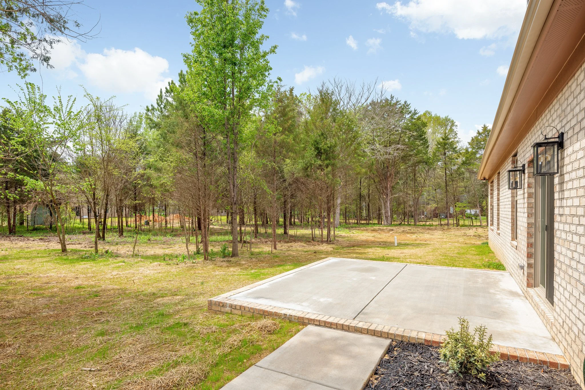 Concrete patio and walkway next to a brick house surrounded by a grassy yard and a wooded area with trees under a blue sky.