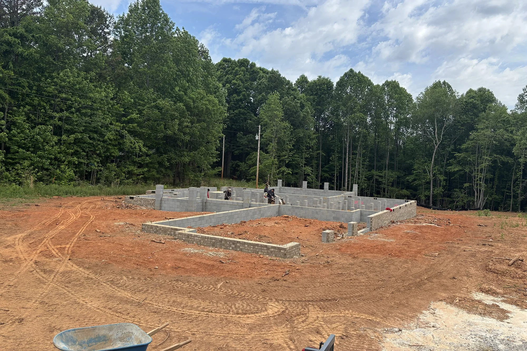 Construction site with concrete block walls being assembled in a wooded area under a partly cloudy sky, with construction workers working and a wheelbarrow in the foreground.