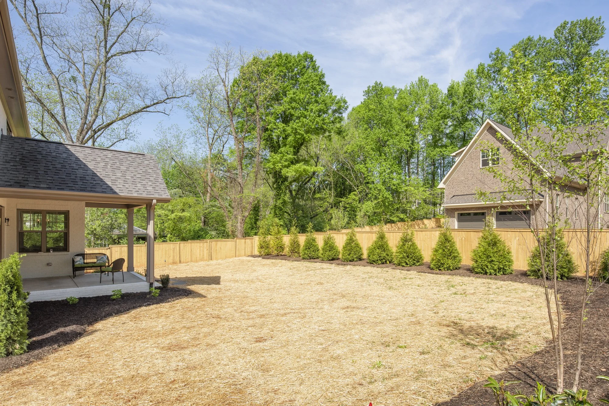 Backyard with brown mulch, young trees along a wooden fence, a patio with chairs, and green trees in the background.