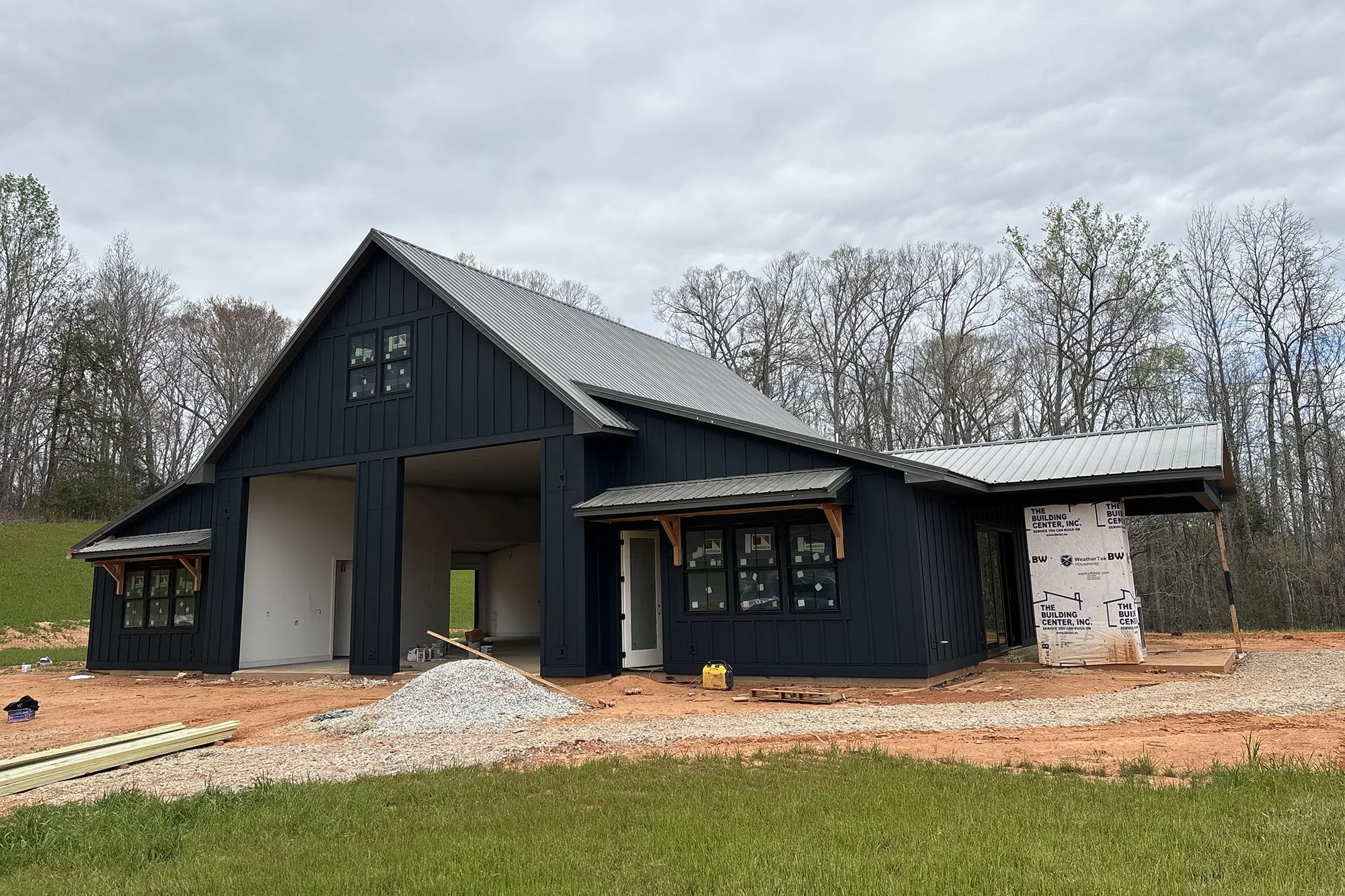 A house under construction with black siding and large windows, surrounded by at least three leafless trees, on a cloudy day with a grassy foreground and construction materials on the ground.