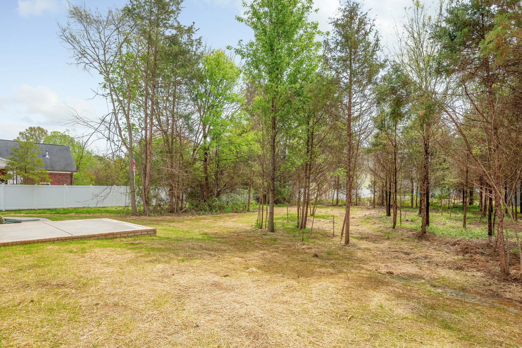 Backyard with patio and trees, grassy area, white fence, and a house in background.