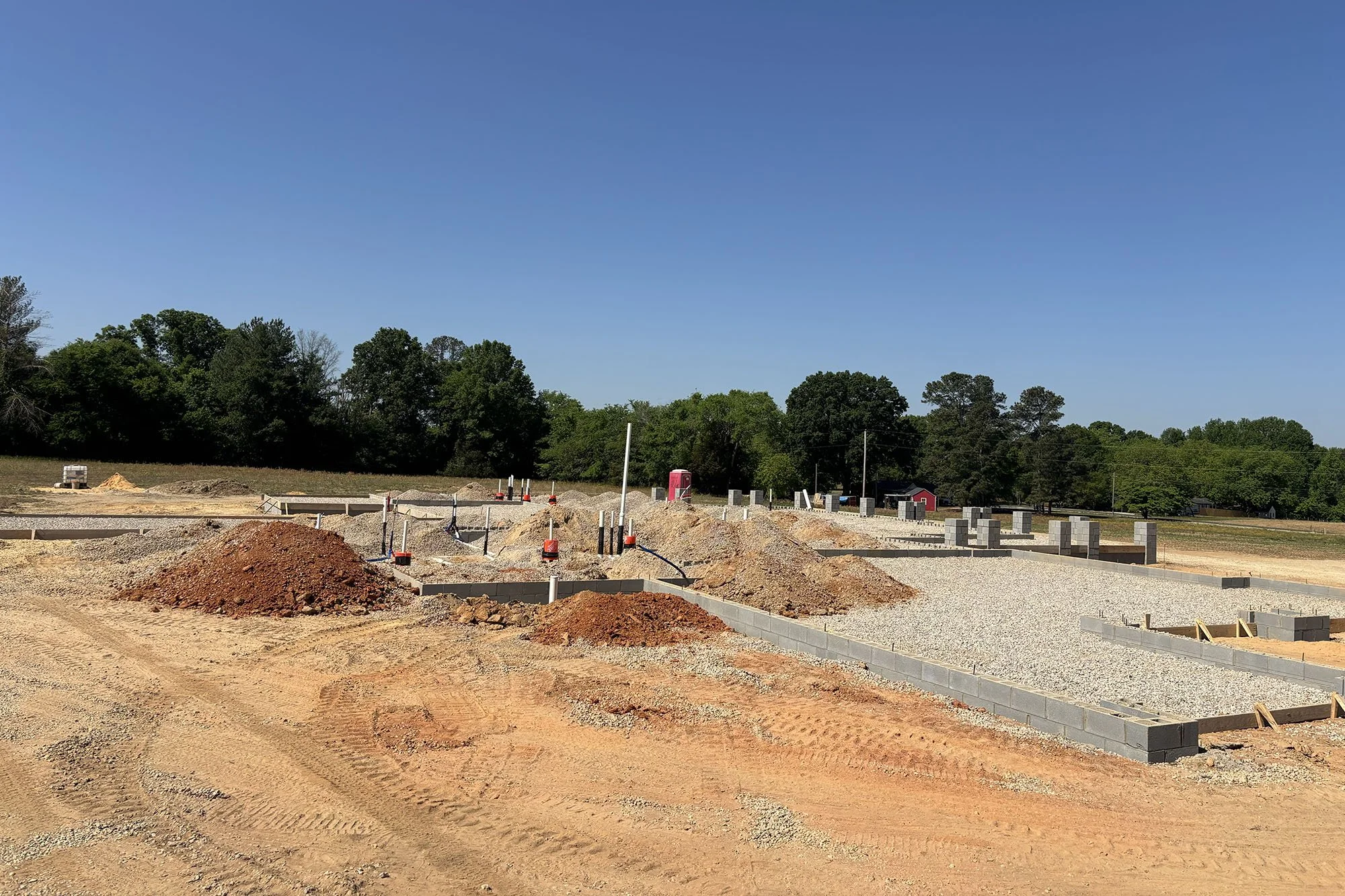 Construction site with gravel and dirt, partially built cinder block walls, orange construction cones, and a wooded area with green trees under a clear blue sky.