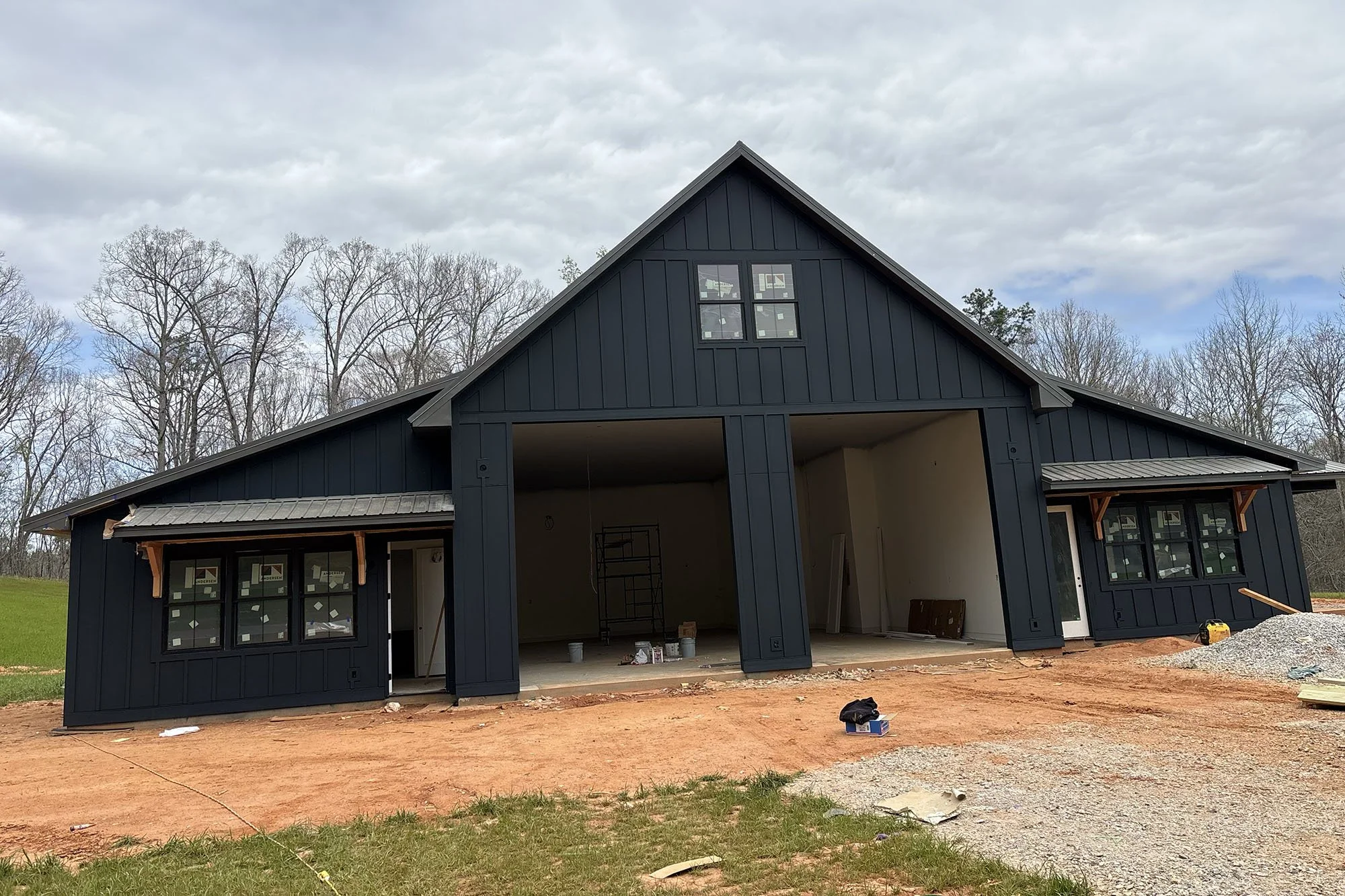 A house under construction with black exterior siding and large windows, situated on a dirt lot with some construction tools and materials nearby, and a background of leafless trees under a cloudy sky.