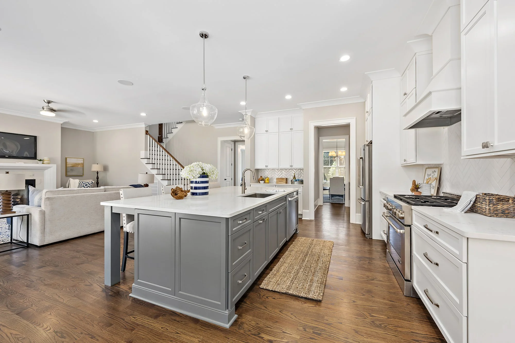 Modern open-plan kitchen and living room with hardwood floors, gray island with marble countertop, white cabinets, stainless steel appliances, pendant lights, and a cozy seating area with a staircase in the background.