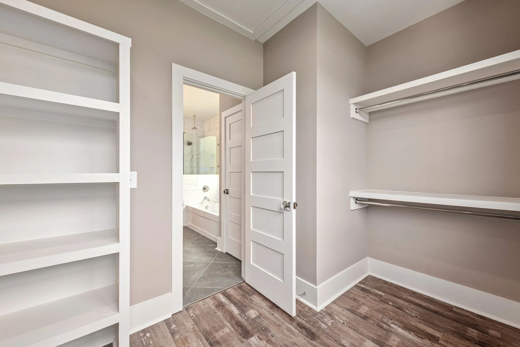 Modern walk-in closet with built-in shelving and hanging rods, leading to a bathroom with a bathtub and tiled flooring.