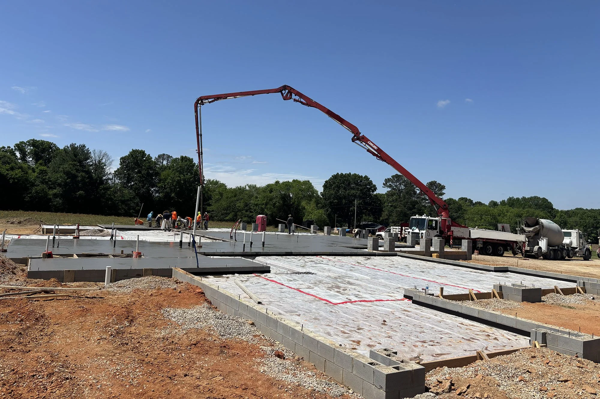 Construction site with workers pouring concrete foundation under a clear blue sky, with a concrete pump truck and various construction materials.