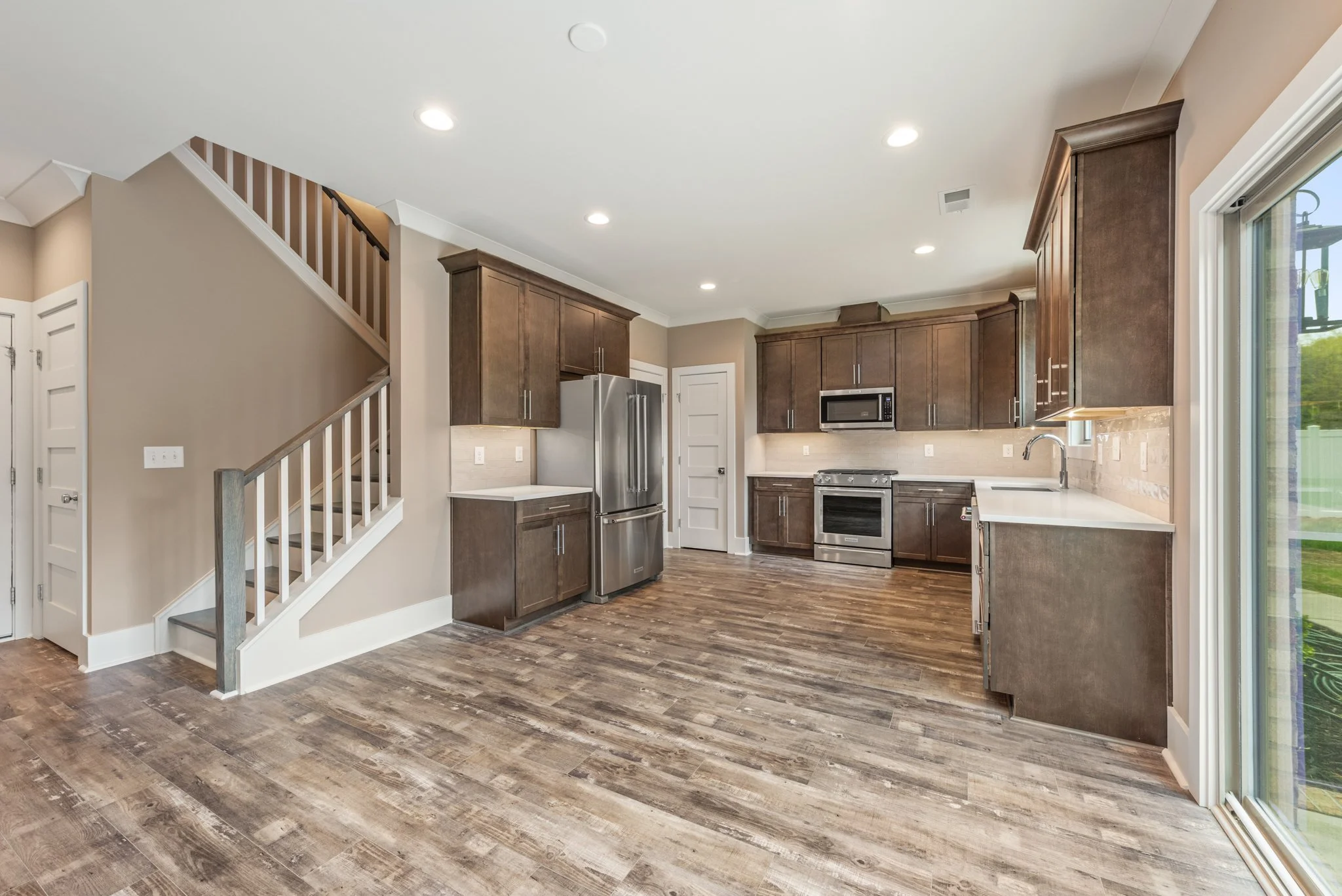 Modern kitchen with wooden cabinets, stainless steel appliances, hardwood flooring, and a staircase. A large window provides natural light.