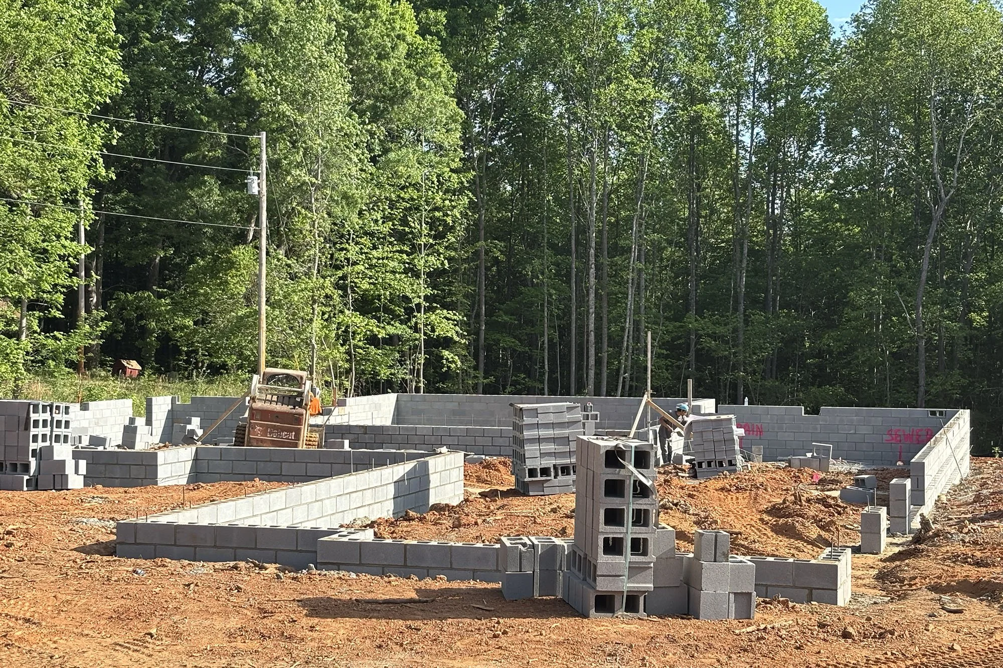 Construction site with cinder block walls being built, construction workers, machinery, and a background of lush green trees.