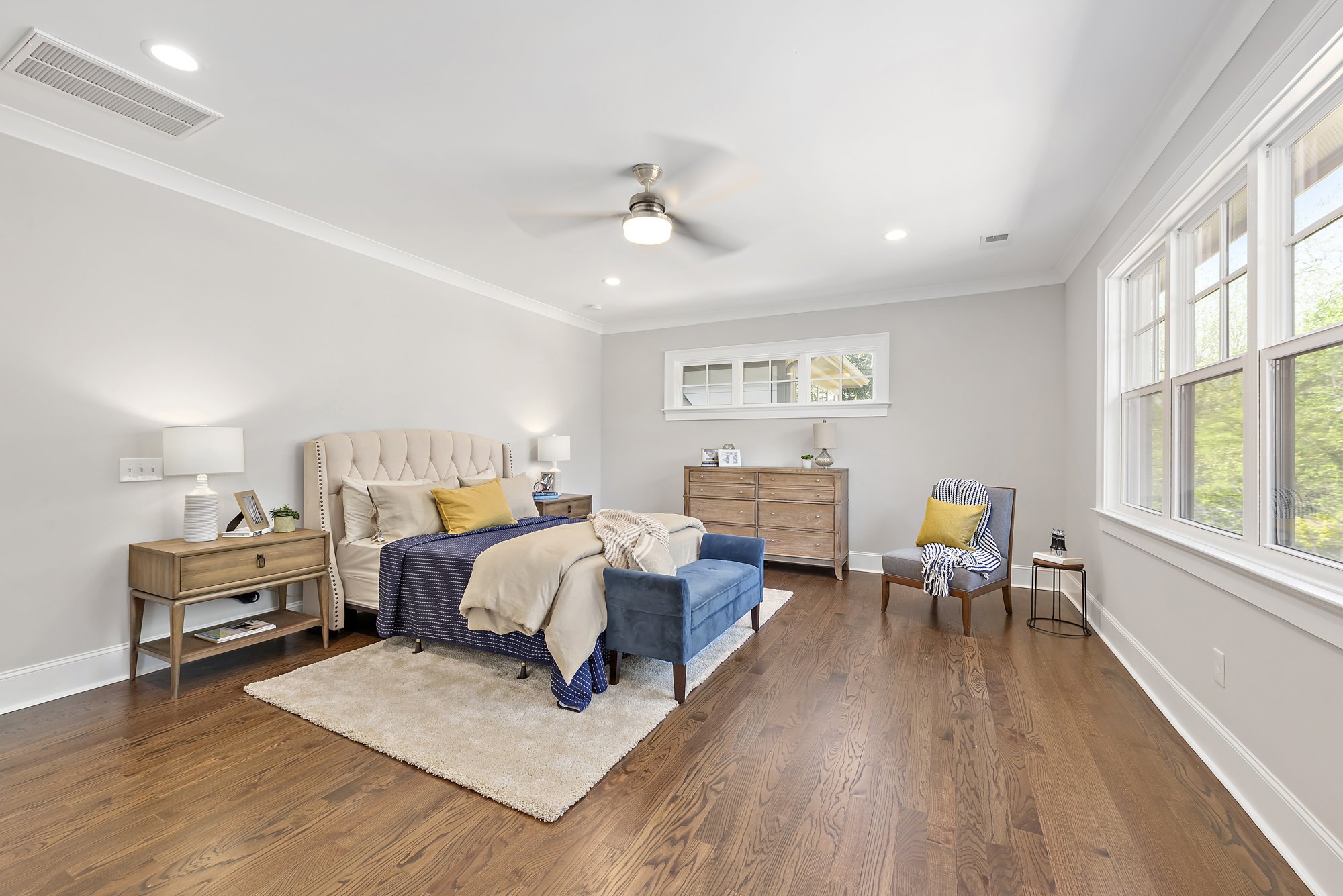Modern bedroom with beige and blue decor, featuring a large bed, nightstands, dresser, armchair, ceiling fan, hardwood floors, and large windows.