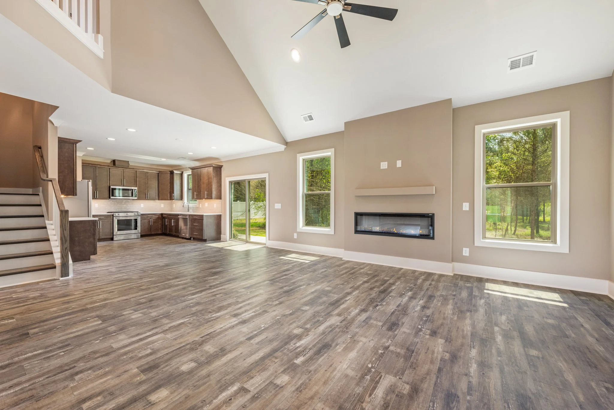 Open-concept living room and kitchen with wooden flooring, brown cabinetry, stainless steel appliances, staircase, fireplace, ceiling fan, and large windows with a view of greenery.