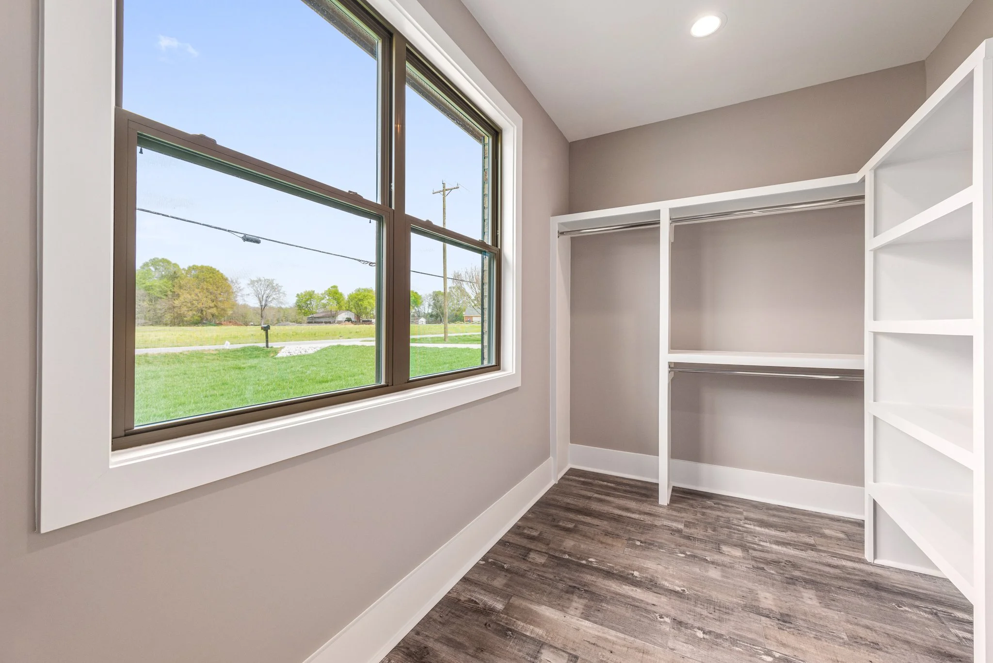 Empty walk-in closet with wooden floor and large window overlooking green grass and trees.