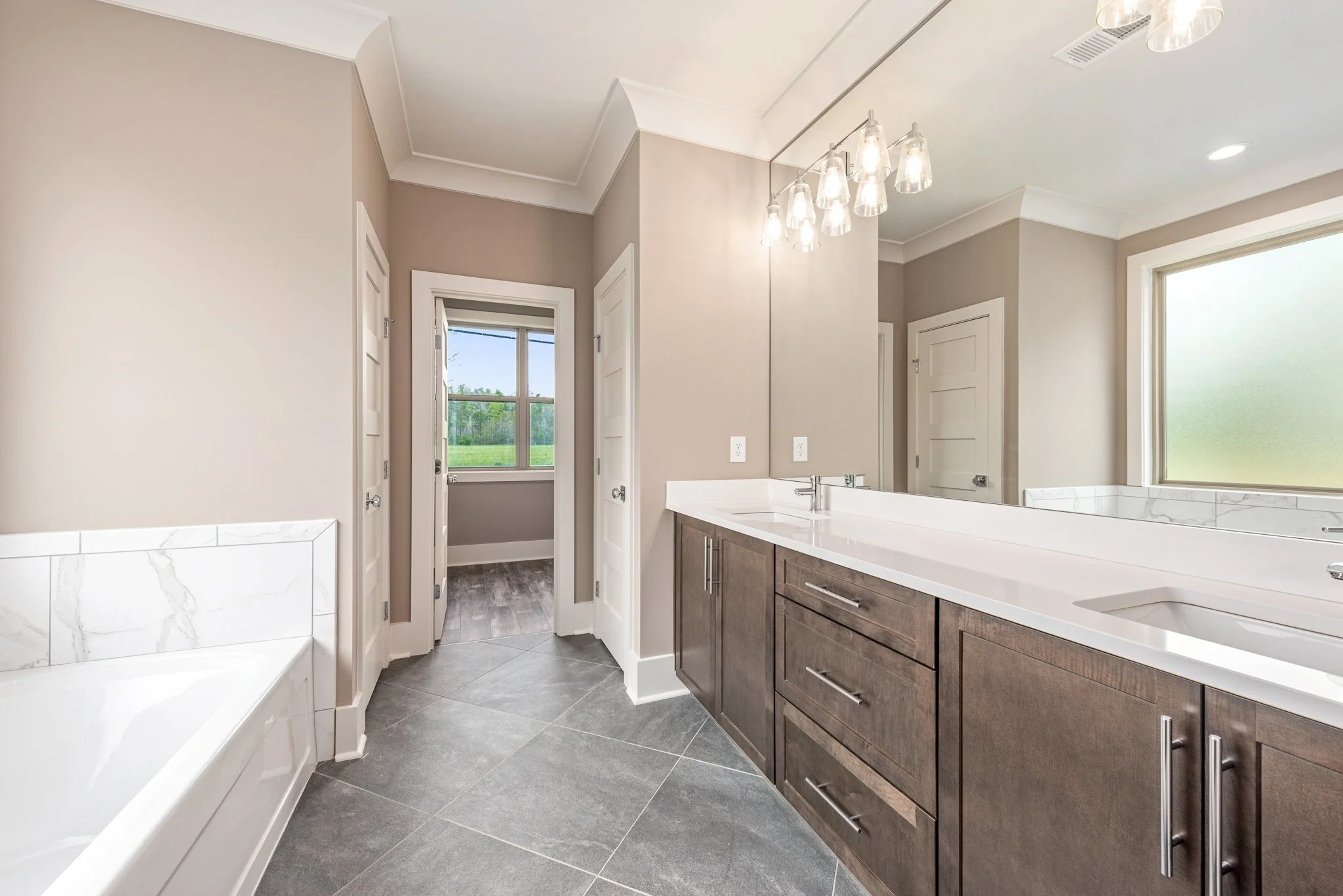 Modern bathroom with large mirror, dual sinks, dark wood vanity, gray tile floor, and a white bathtub. There is a doorway leading to another room with a window.