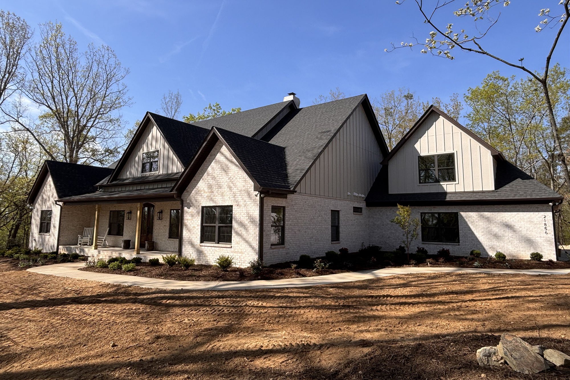 A modern house with white brick and wood siding exterior, multiple gabled roofs, and a small porch with rocking chairs. The house is surrounded by trees and has a cleared, dirt front yard.