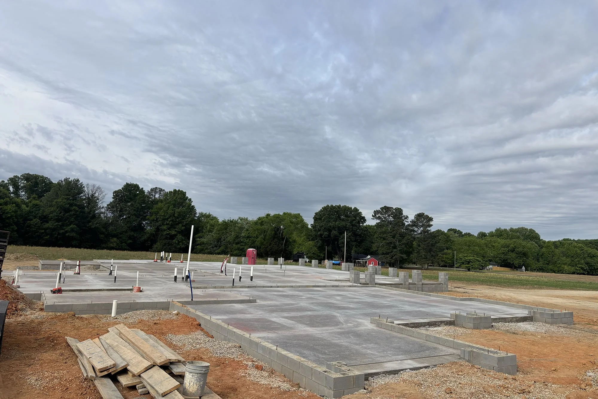 Construction site with concrete foundation and steel reinforcements, surrounded by trees and cloudy sky.