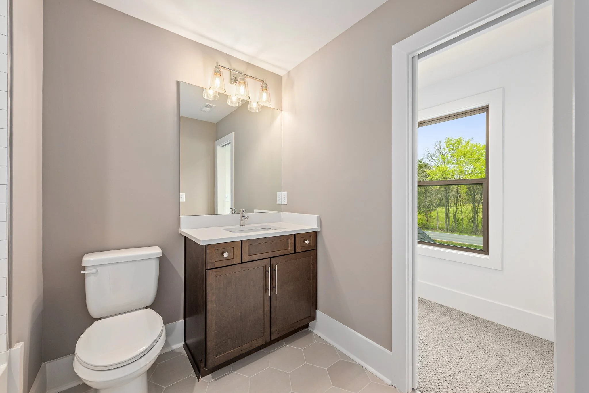 Modern bathroom with a toilet, wooden vanity, large mirror, and a three-light fixture, adjacent to a room with a window showing greenery outside.
