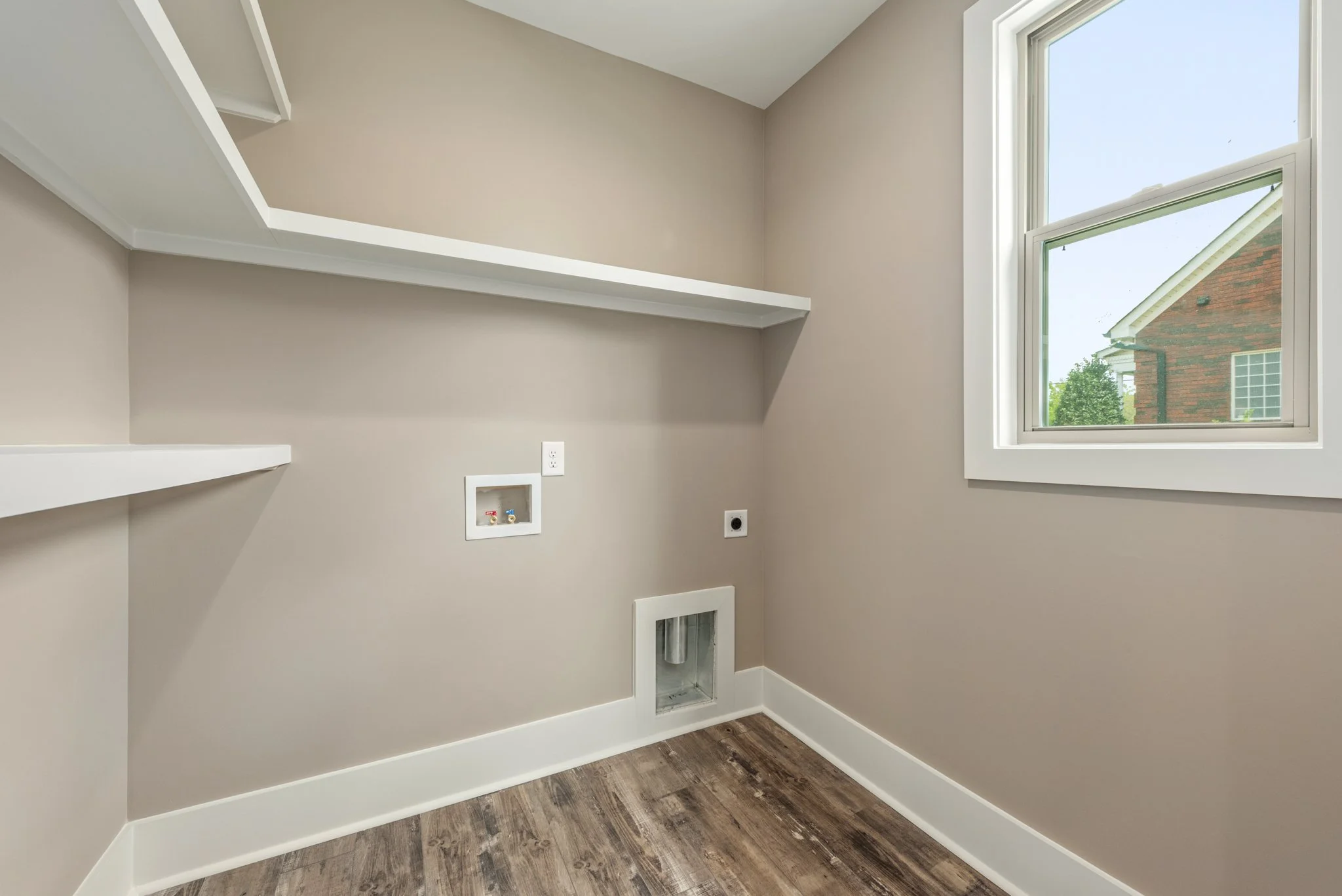 Empty laundry room with beige walls, white trim, and wood flooring, featuring a window, utility connections, and upper shelves.