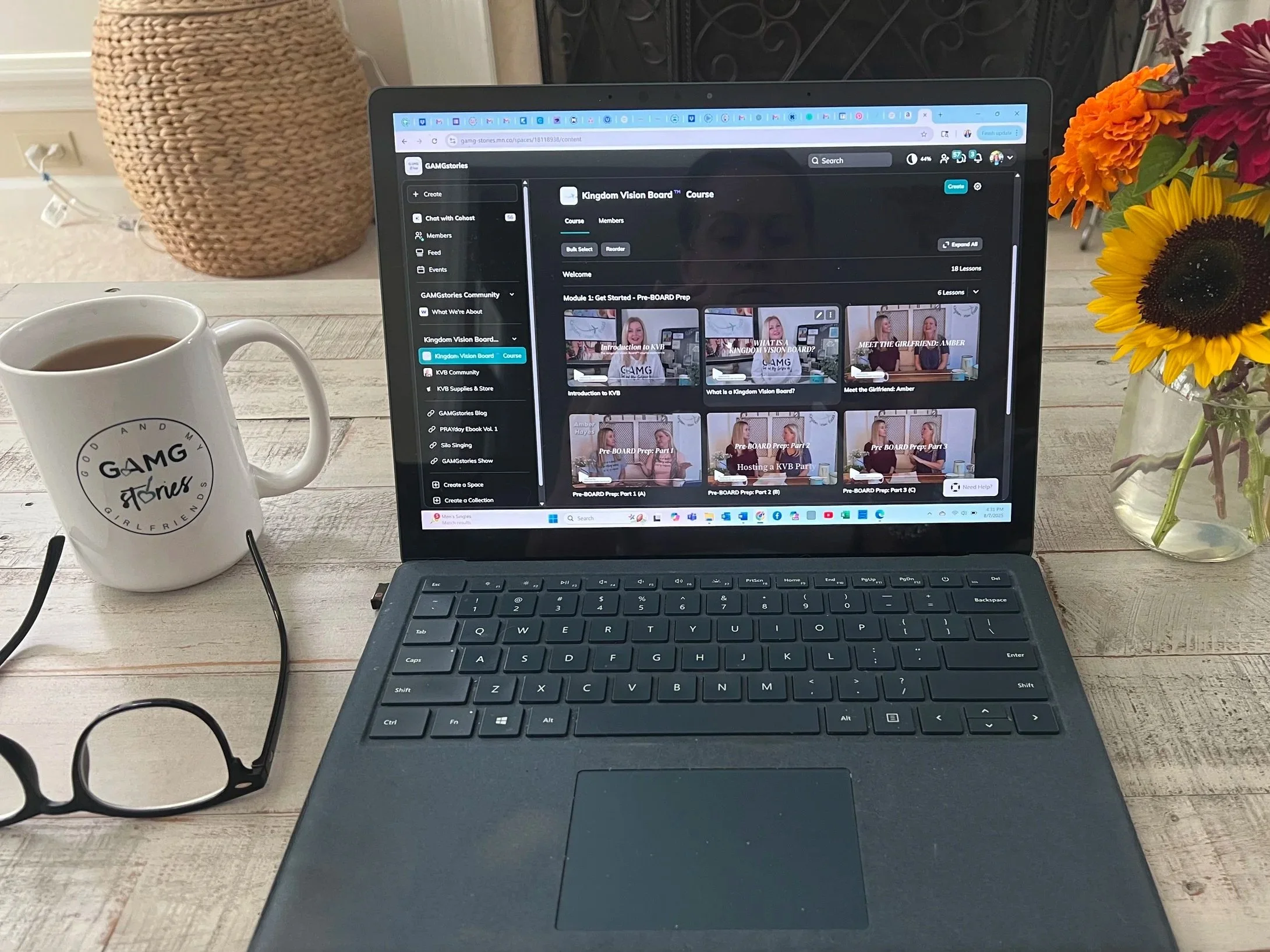 A laptop on a wooden table showing an online course titled 'Kingdom Vision Board Course' with a coffee mug, a pair of glasses, a woven basket, and a vase of colorful flowers nearby.