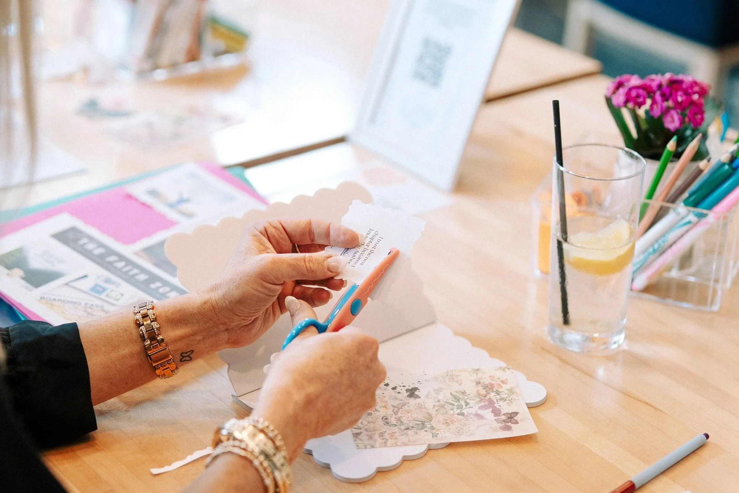 Person cutting decorative paper with scissors at a craft table with floral paper, markers, and a glass of lemonade with lemon slices.