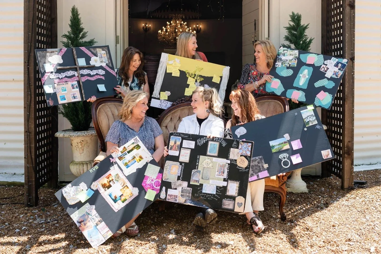 Group of women with large display boards sharing stories and memories, seated and standing outdoors in front of a house with string lights and a chandelier inside.