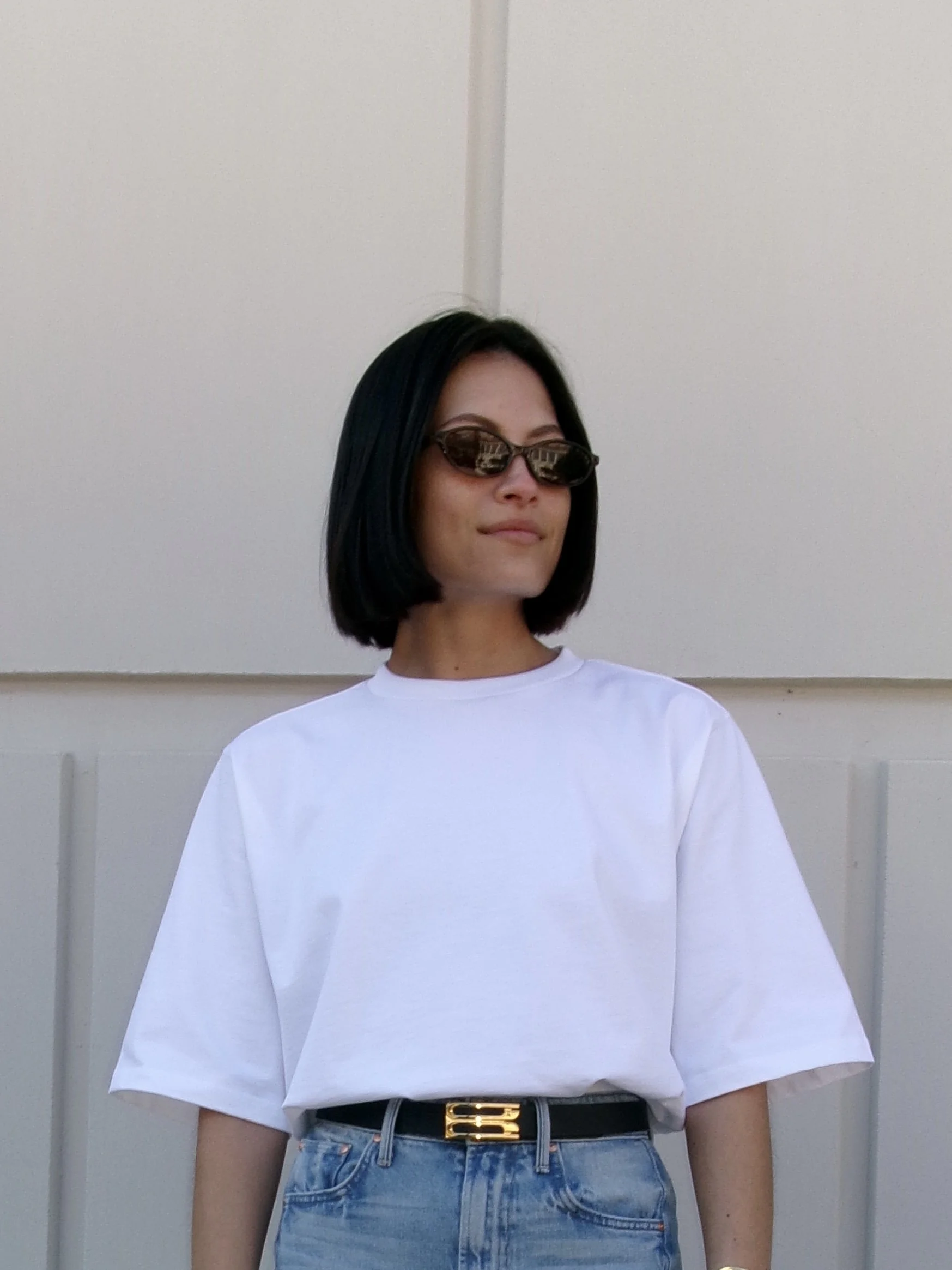 Woman with shoulder-length black hair wearing sunglasses, white t-shirt, and blue jeans with a black belt, standing in front of a plain white wall.