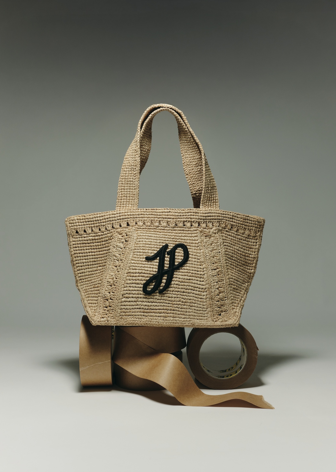 A woven beige handbag with black embroidered initials 'JP' on the front, placed on rolls of brown packing tape and paper.