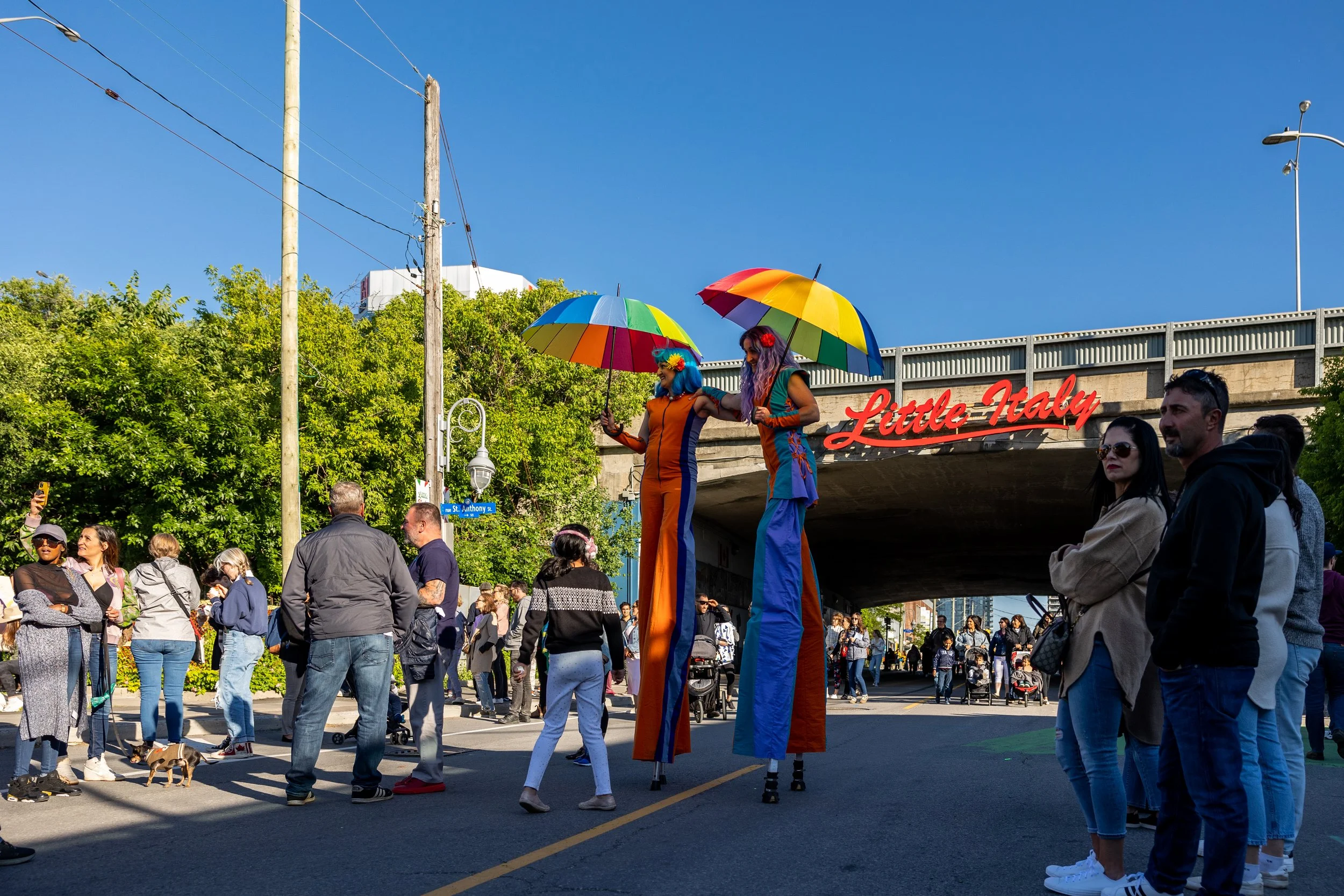 Street scene in Little Italy with two stilt walkers holding rainbow umbrellas, surrounded by a crowd of people under a clear blue sky.