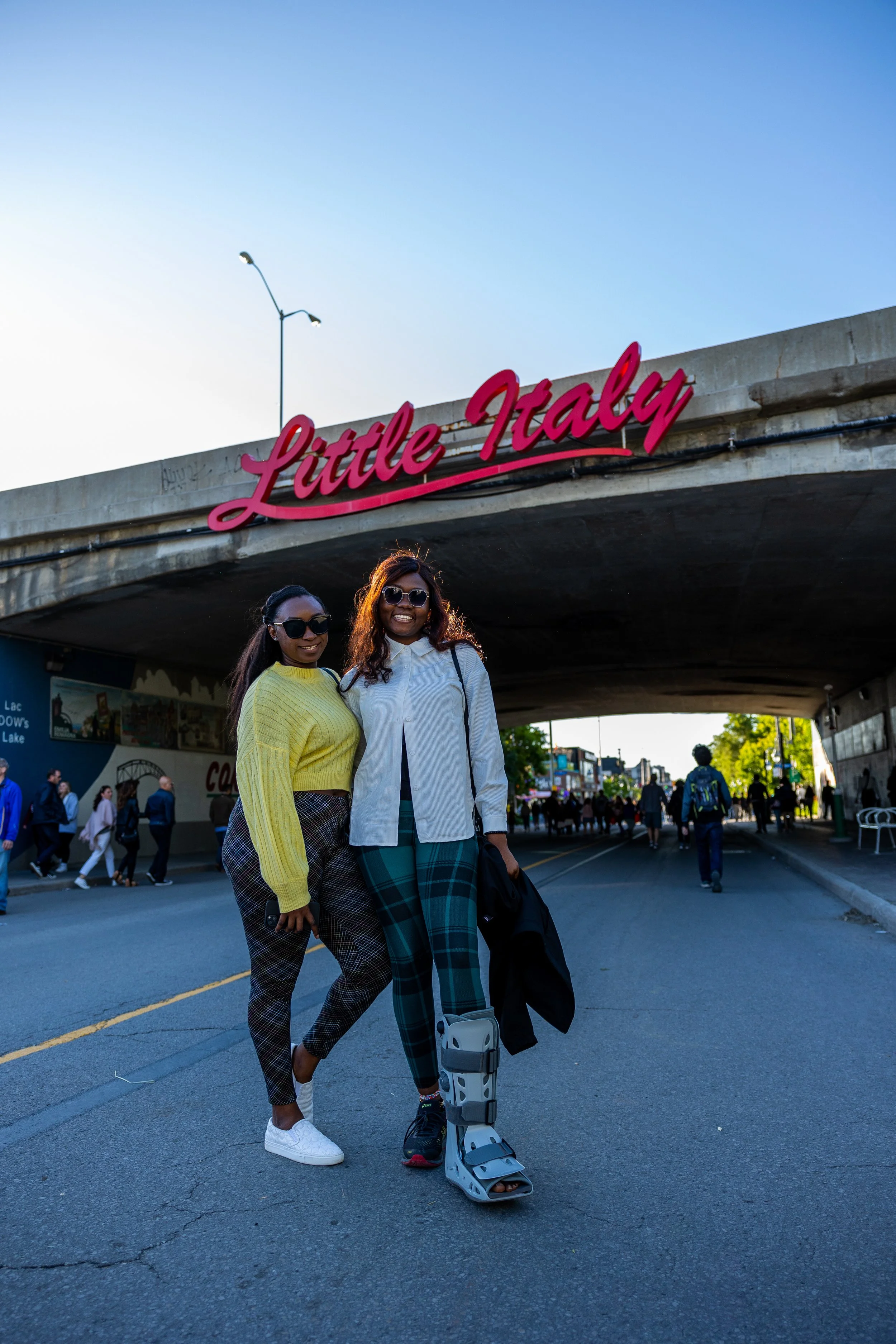 Two people posing under "Little Italy" sign, one wearing a walking boot.