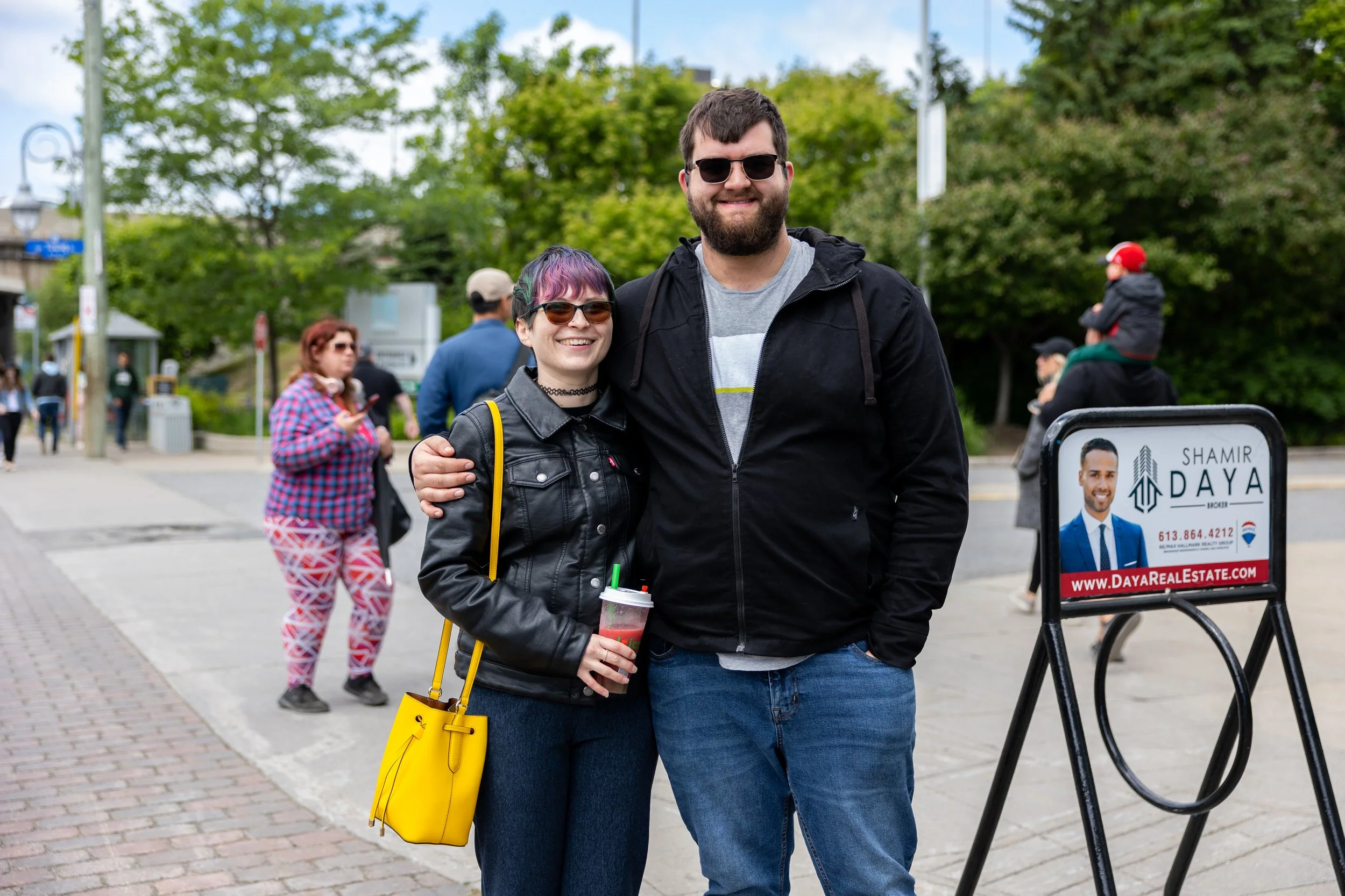 A couple poses for a photo on a sidewalk. The woman, with a colorful short hairstyle, wears sunglasses, a black leather jacket, and holds a yellow bag and a drink. The man, also in sunglasses, wears a black hoodie. Other people and trees are visible 