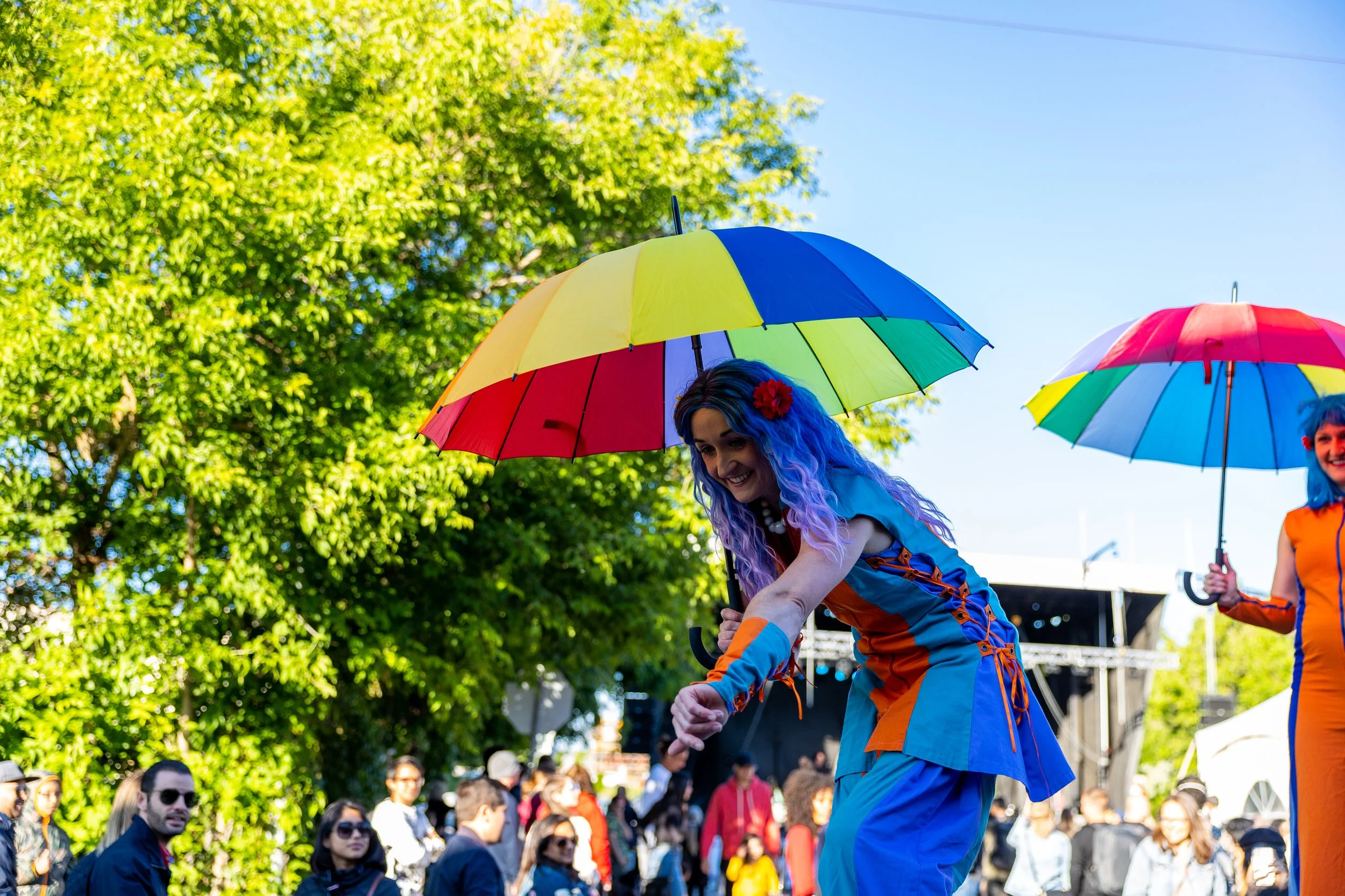 Performers with colorful umbrellas at an outdoor event, surrounded by a crowd and trees.