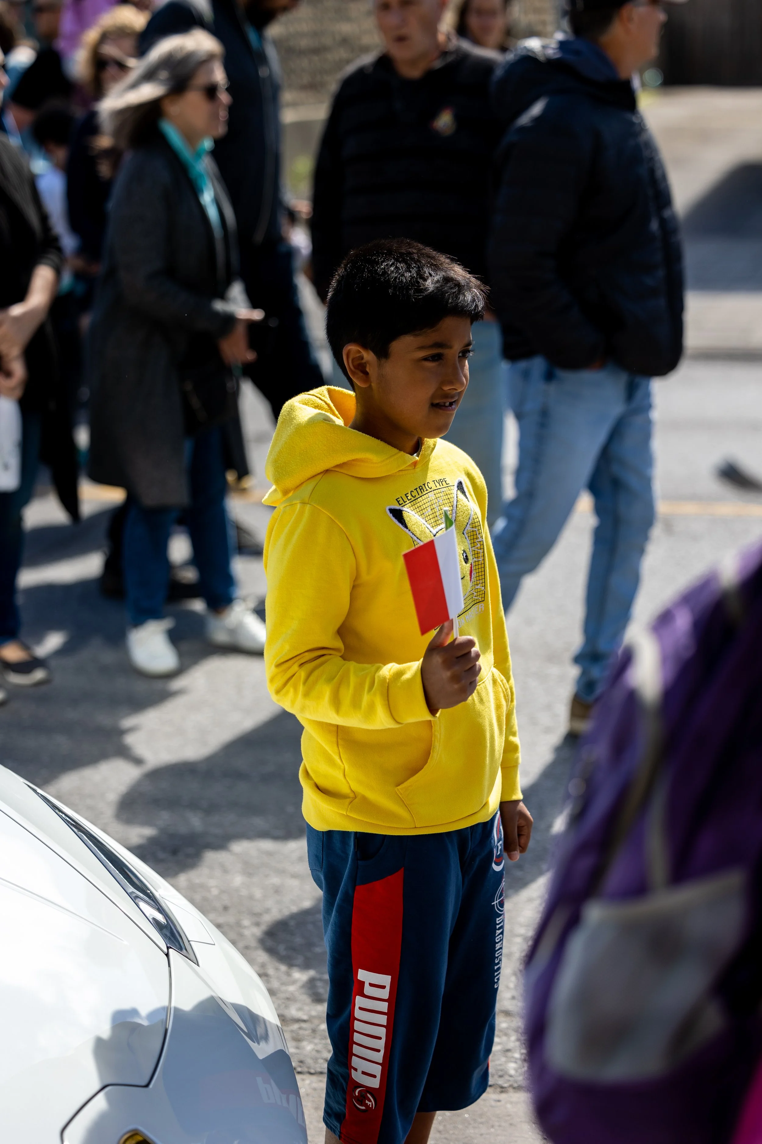 A boy wearing a yellow hoodie and shorts holds a small flag among a crowd of people on a street.