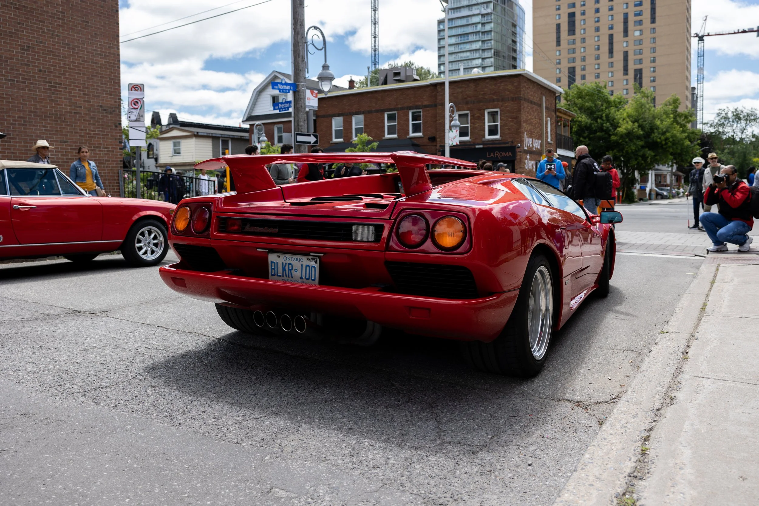 Red Lamborghini Diablo parked on a city street with pedestrians and another vintage red car nearby.