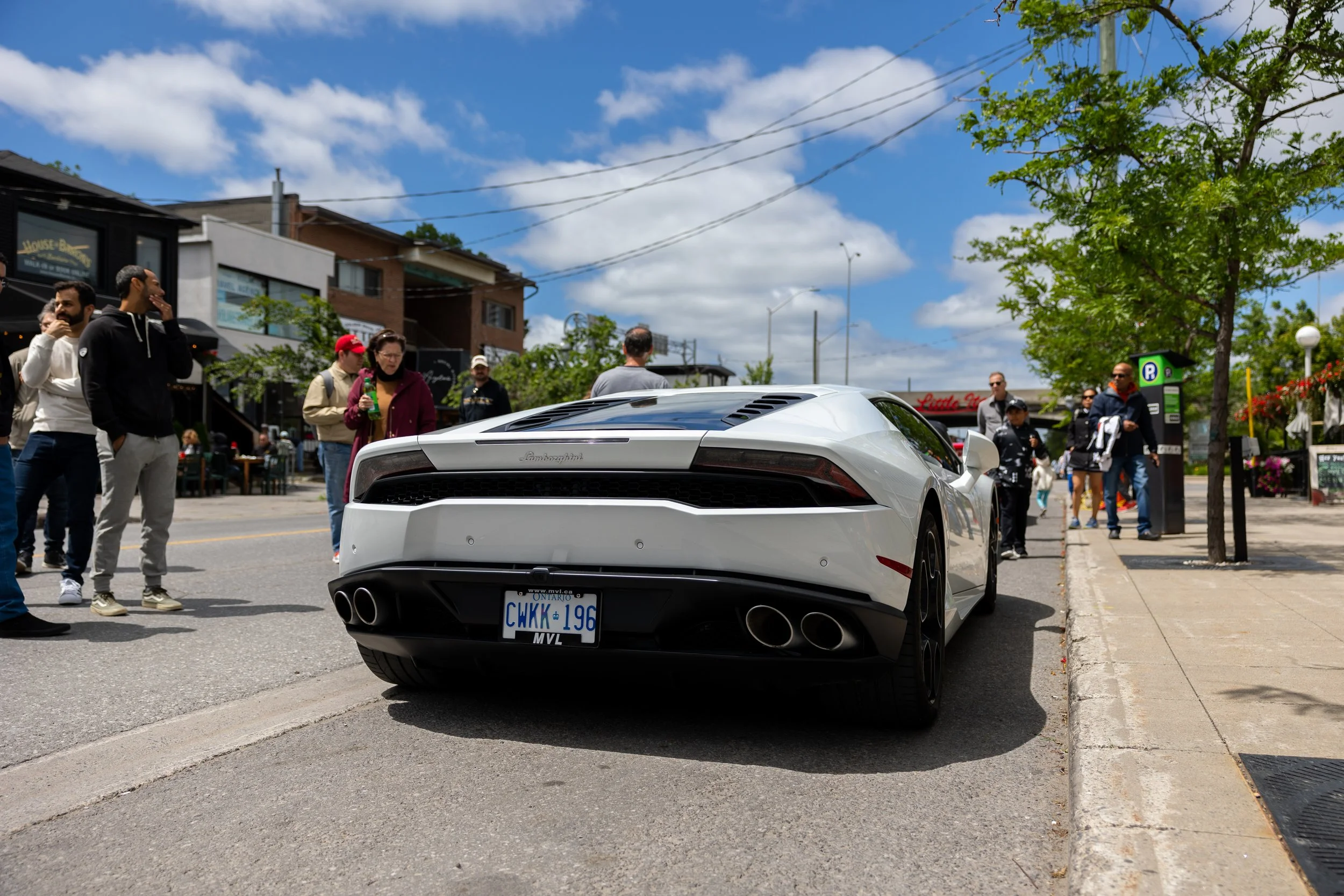 Rear view of a white Lamborghini sports car parked on a street, with people standing nearby observing the car and partially cloudy sky above.