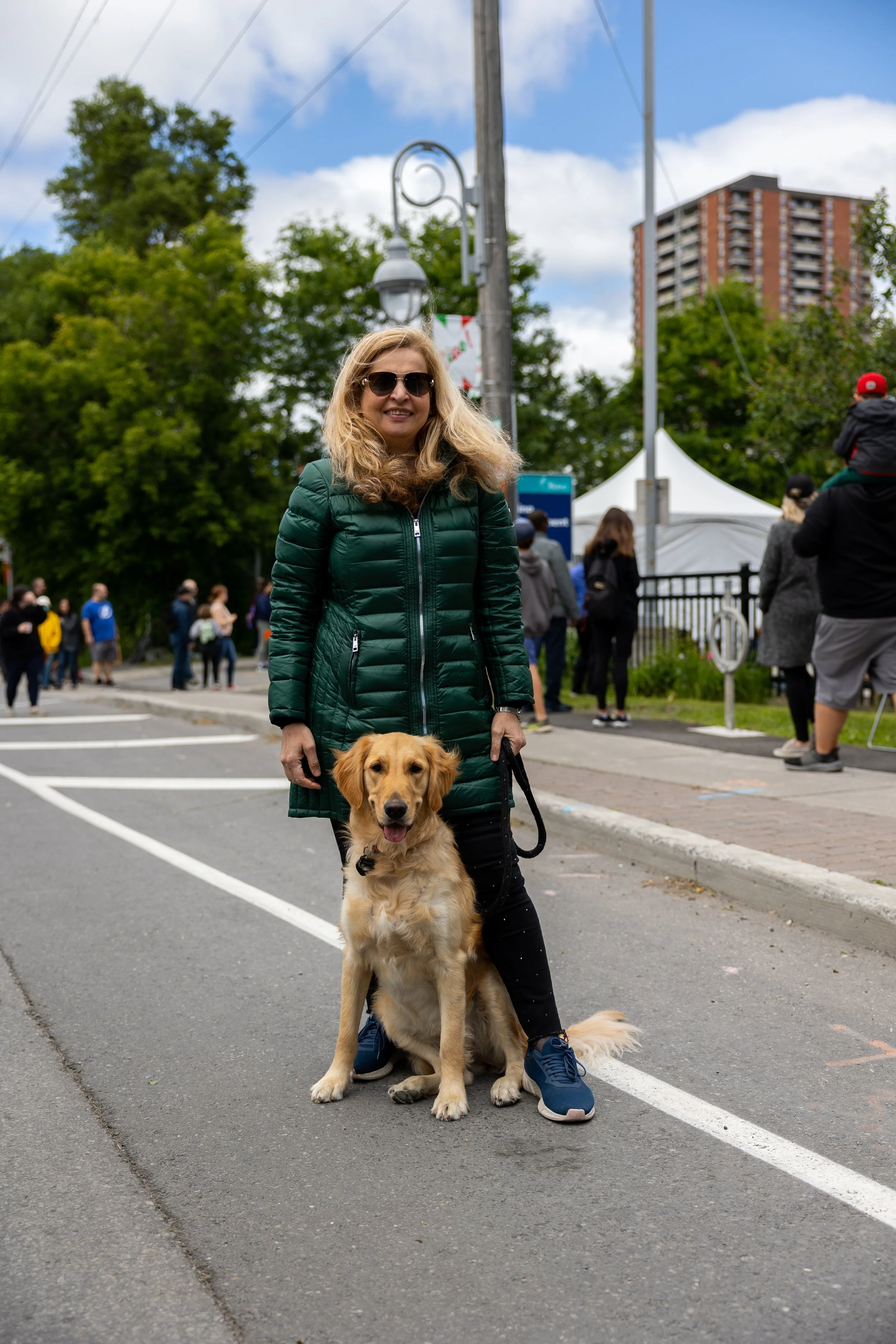Woman in green coat with sunglasses standing on a street holding a leash of a golden retriever, with people and trees in the background.