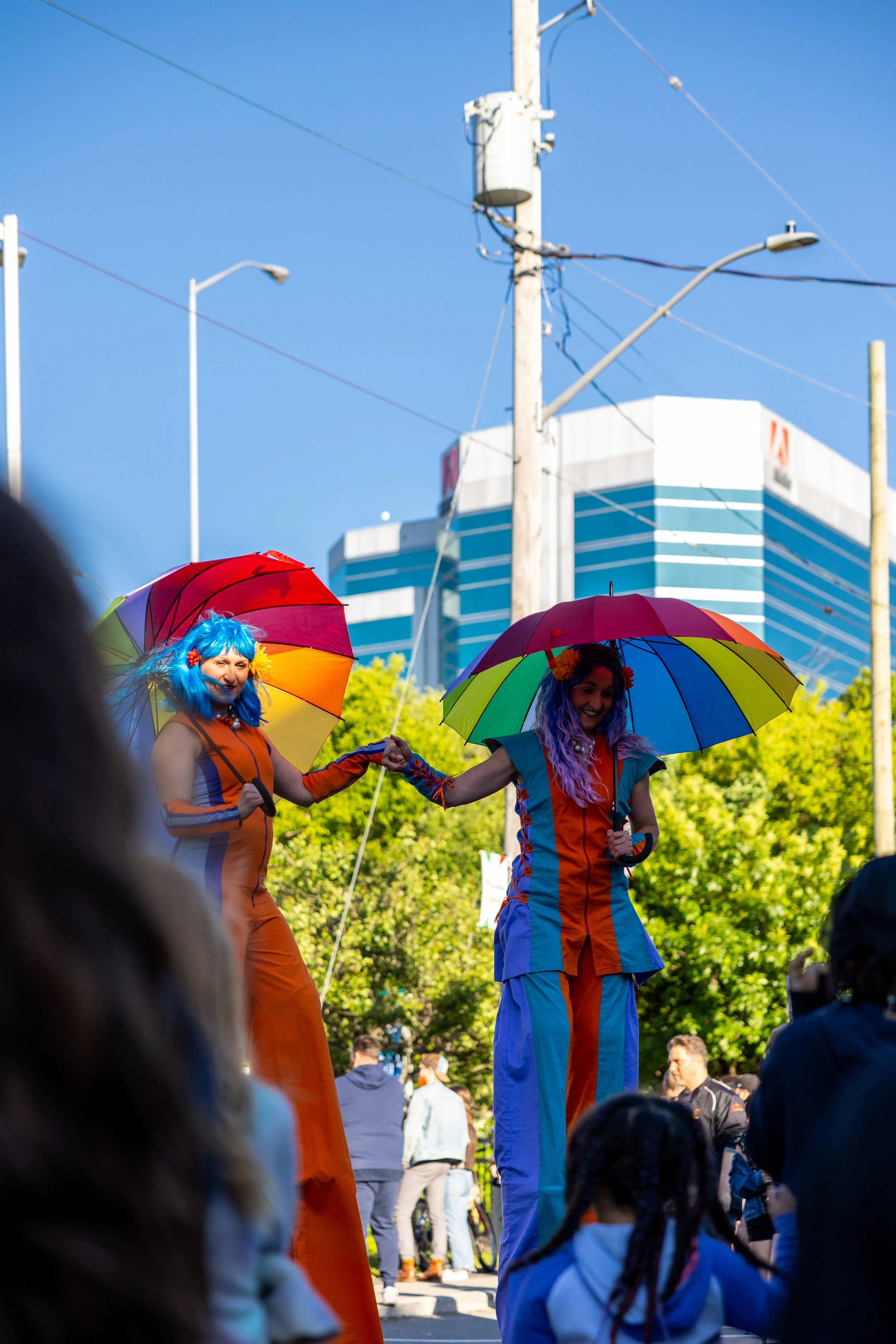 Two performers on stilts wearing colorful costumes and wigs, holding rainbow umbrellas, in a parade or festival setting with a crowd around them.