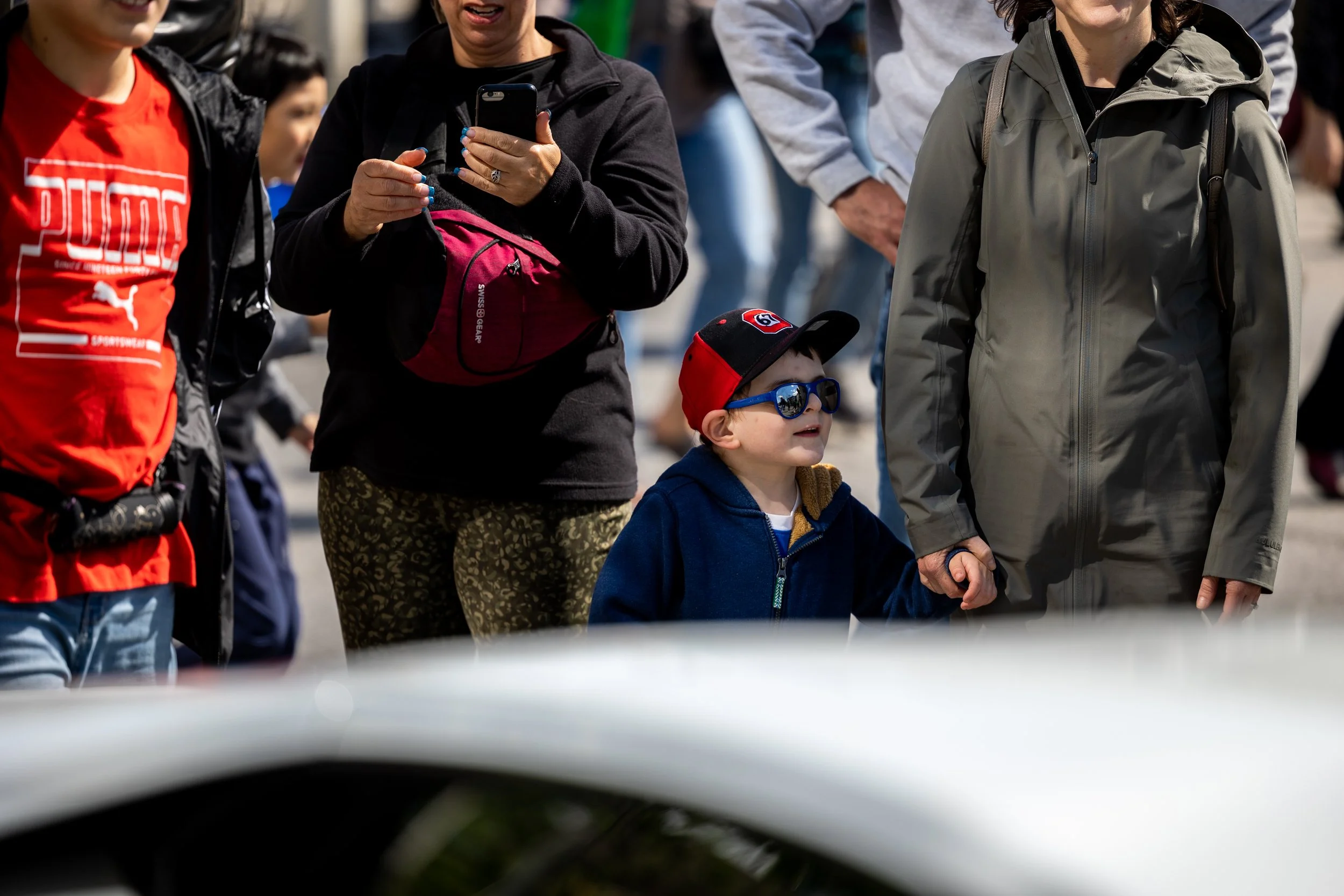 Crowd of people walking outdoors, including a child wearing sunglasses and a cap, holding an adult's hand. Other people are using smartphones and wearing casual clothing.