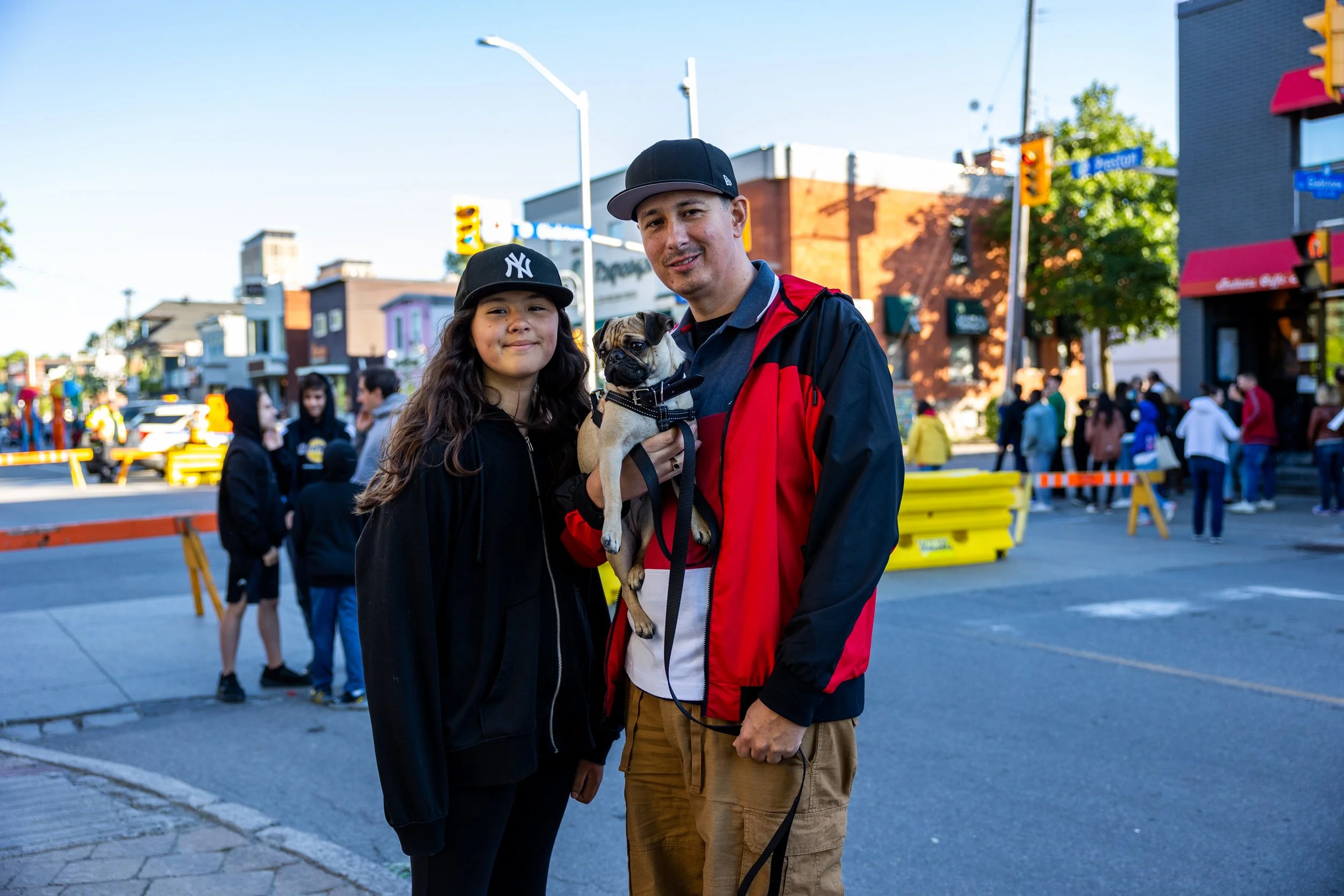 A man and a young girl wearing baseball caps are standing in a street, with the man holding a pug dog. They seem to be at an outdoor event with people in the background. The street is partially blocked by barricades.