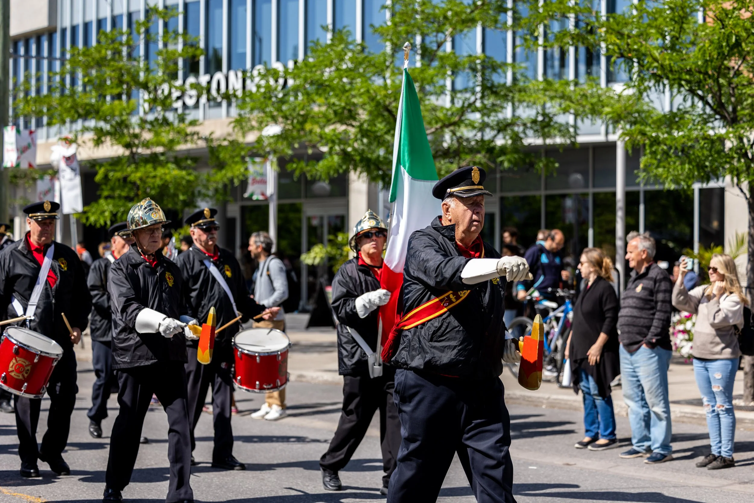 People marching in a parade, some carrying an Italian flag and playing drums, dressed in uniforms, with spectators watching on the sidewalk, and trees and buildings in the background.