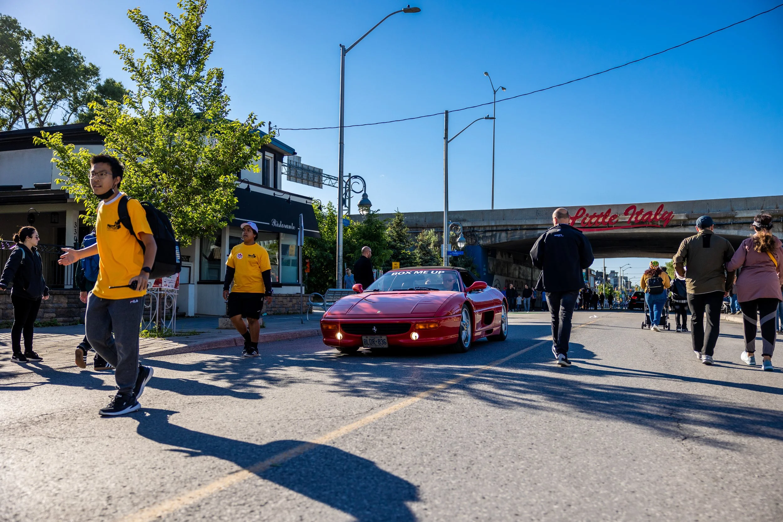 Street scene with a red sports car driving through, people walking around, and a 'Little Italy' sign on an overpass in the background.