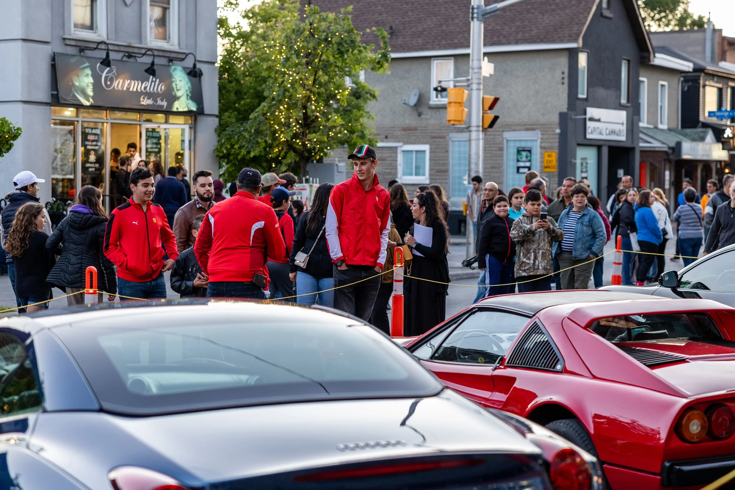 A street scene with a crowd gathered in front of a store named 'Carmelito'. People are wearing casual jackets and some have Ferrari branding. Two sports cars, a red Ferrari and a silver Maserati, are parked in the foreground. The area is cordoned off