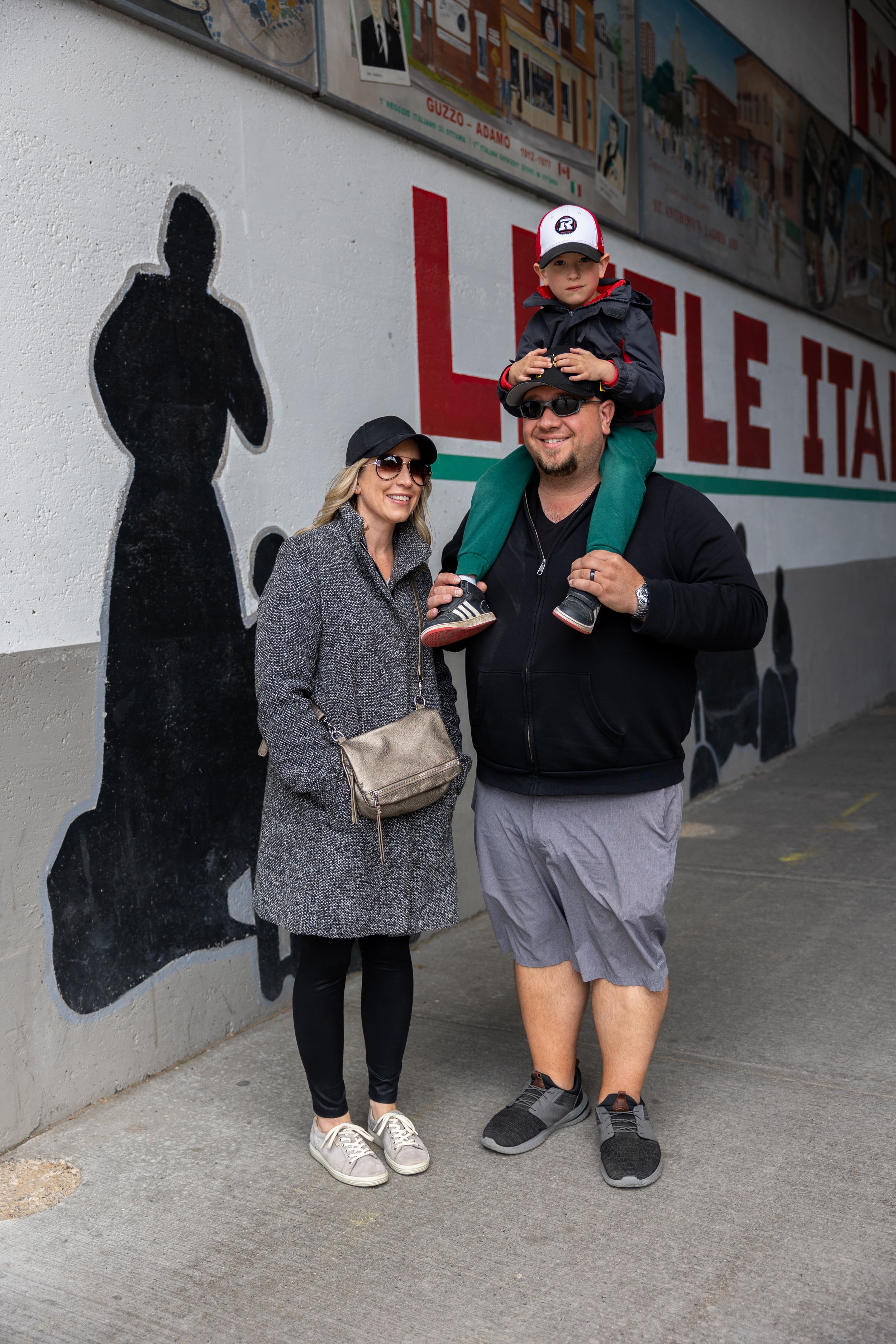 A smiling family standing in front of a mural in Little Italy. A man holds a child on his shoulders, and a woman stands beside them. They are all wearing casual clothing and hats. The mural features black silhouettes and part of the text "LITTLE ITAL