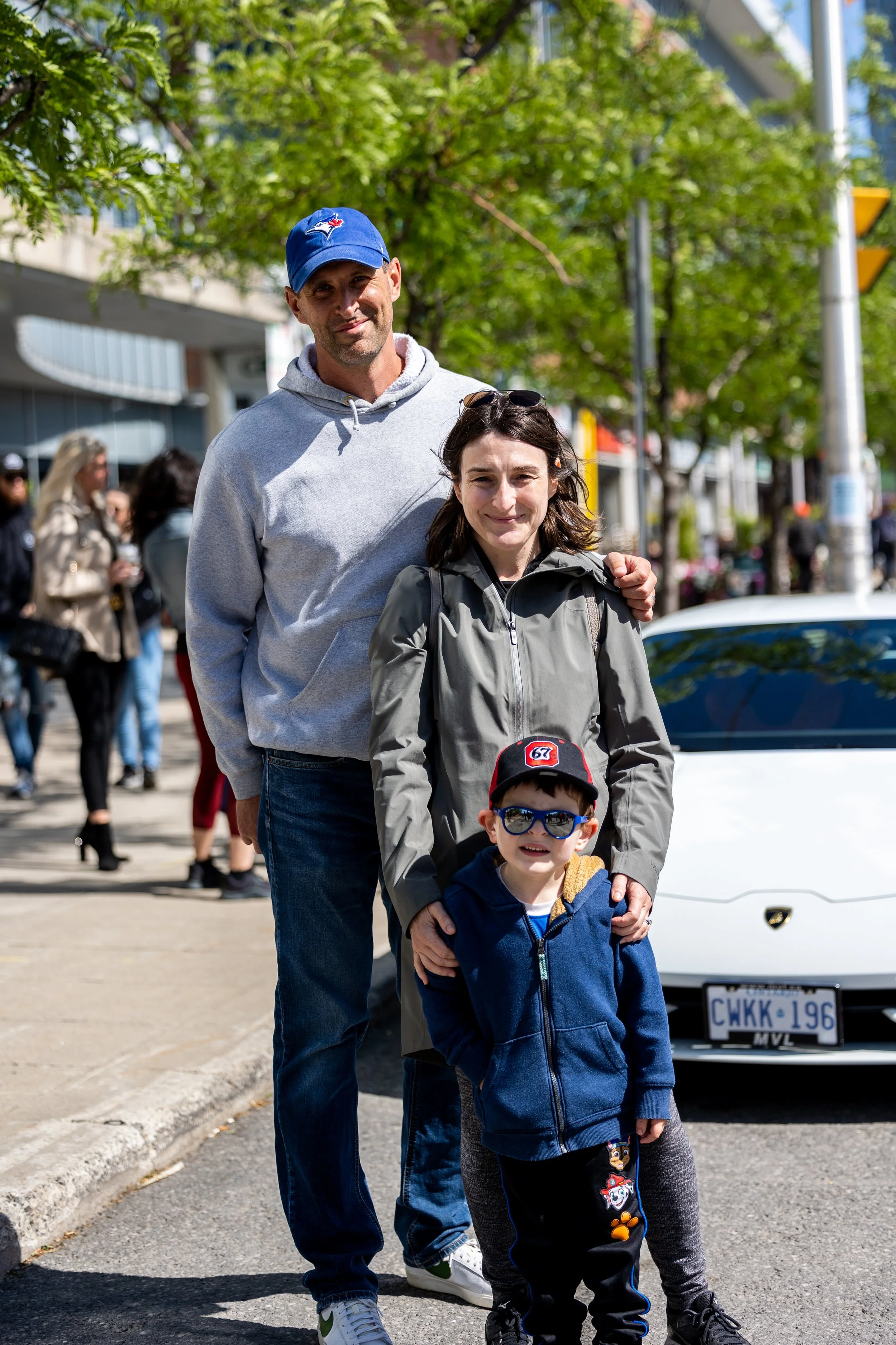 Family posing outdoors with a white sports car in the background and people walking on the sidewalk.